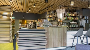 A bar area with wooden slats on the ceiling, a counter with a stack of books, and shelves with various bottles and glasses.