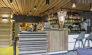 A bar area with wooden slats on the ceiling, a counter with a stack of books, and shelves with various bottles and glasses.