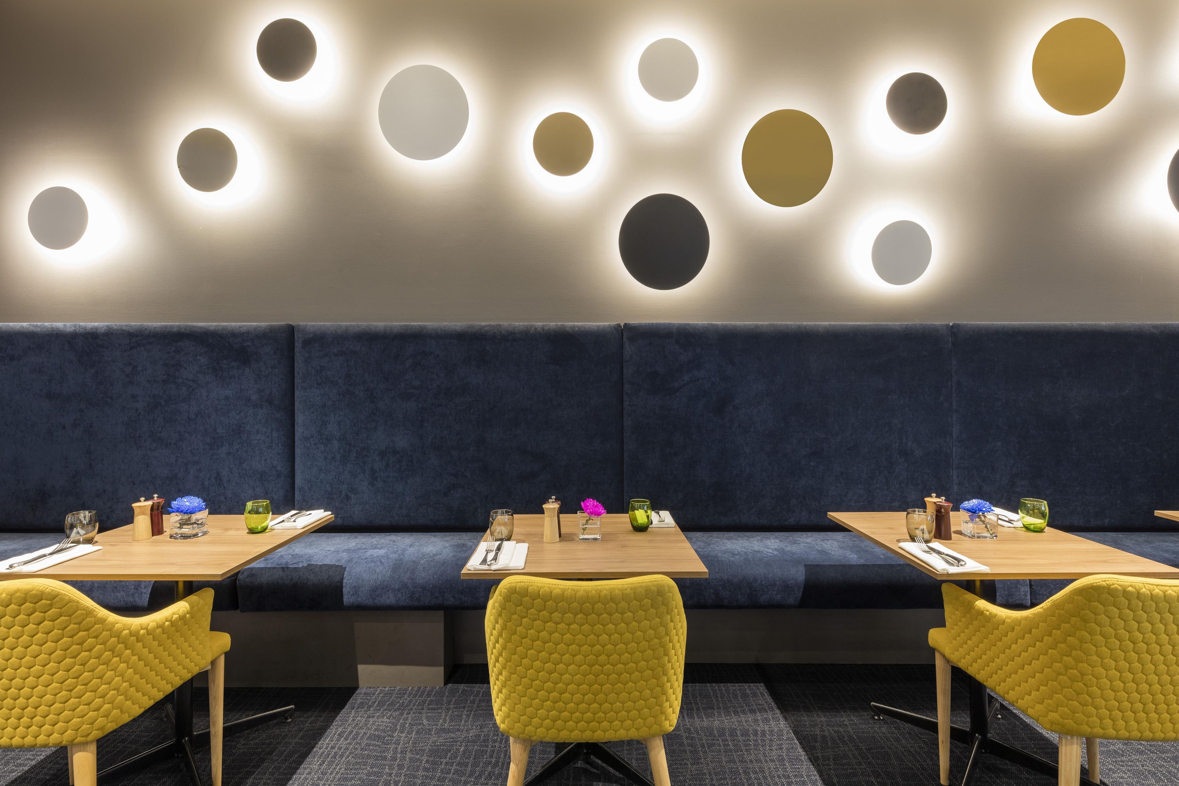 A modern restaurant with a dark blue cushioned bench seating, wooden tables, and yellow chairs.