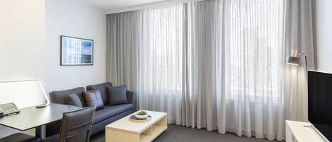 Una habitación de hotel moderna con un sofá gris, una pequeña mesa de centro de madera, un televisor, un escritorio con una silla y un gran ventanal con cortinas traslúcidas.