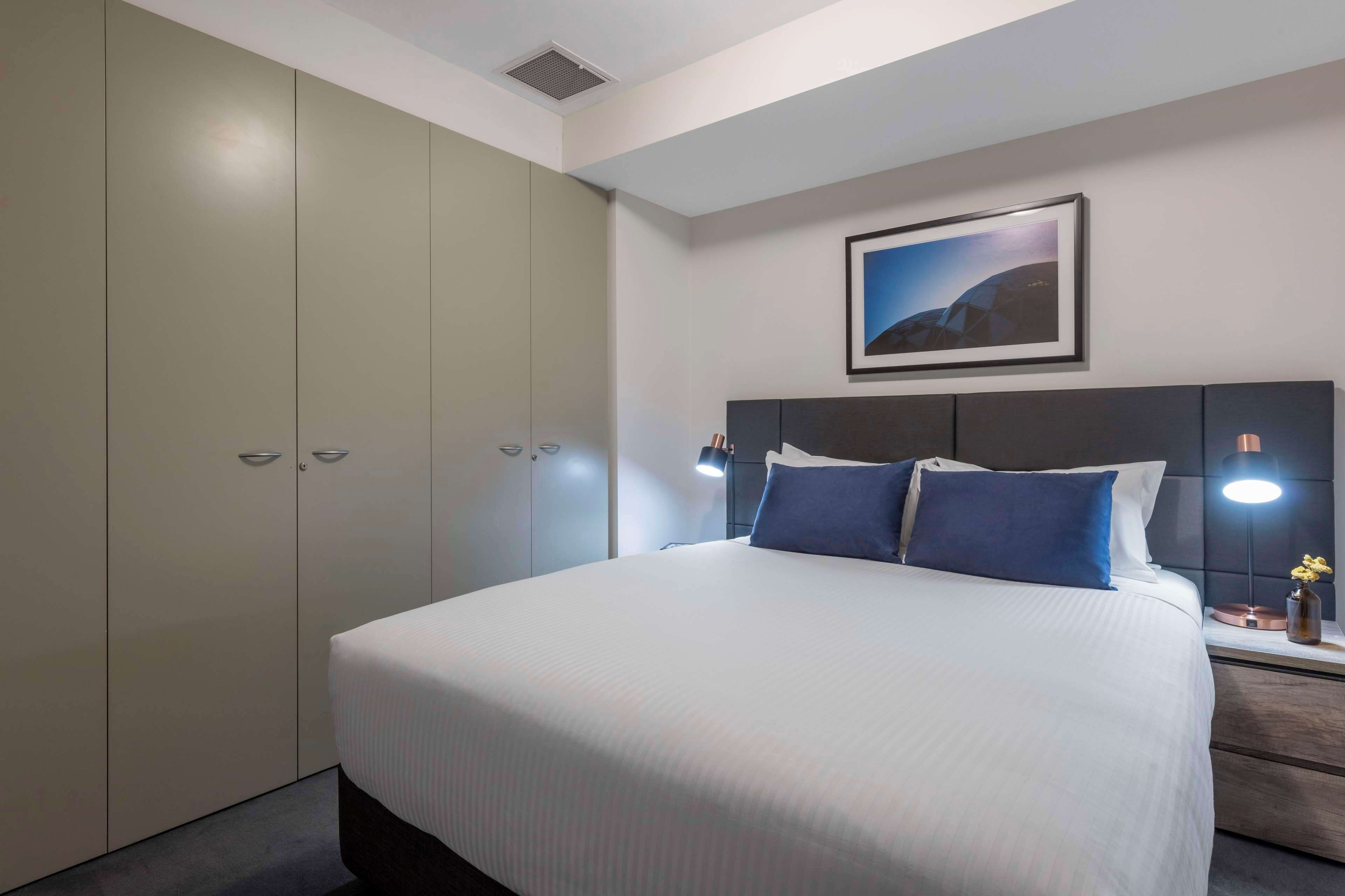 Quarto de hotel com cama grande, cabeceira com luz, mesa de cabeceira com abajur e guarda-roupa.