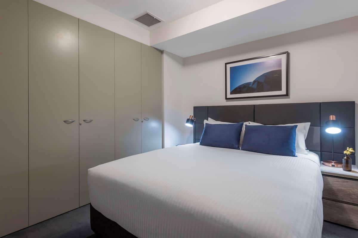 Quarto de hotel com cama grande, cabeceira com luz, mesa de cabeceira com abajur e guarda-roupa.