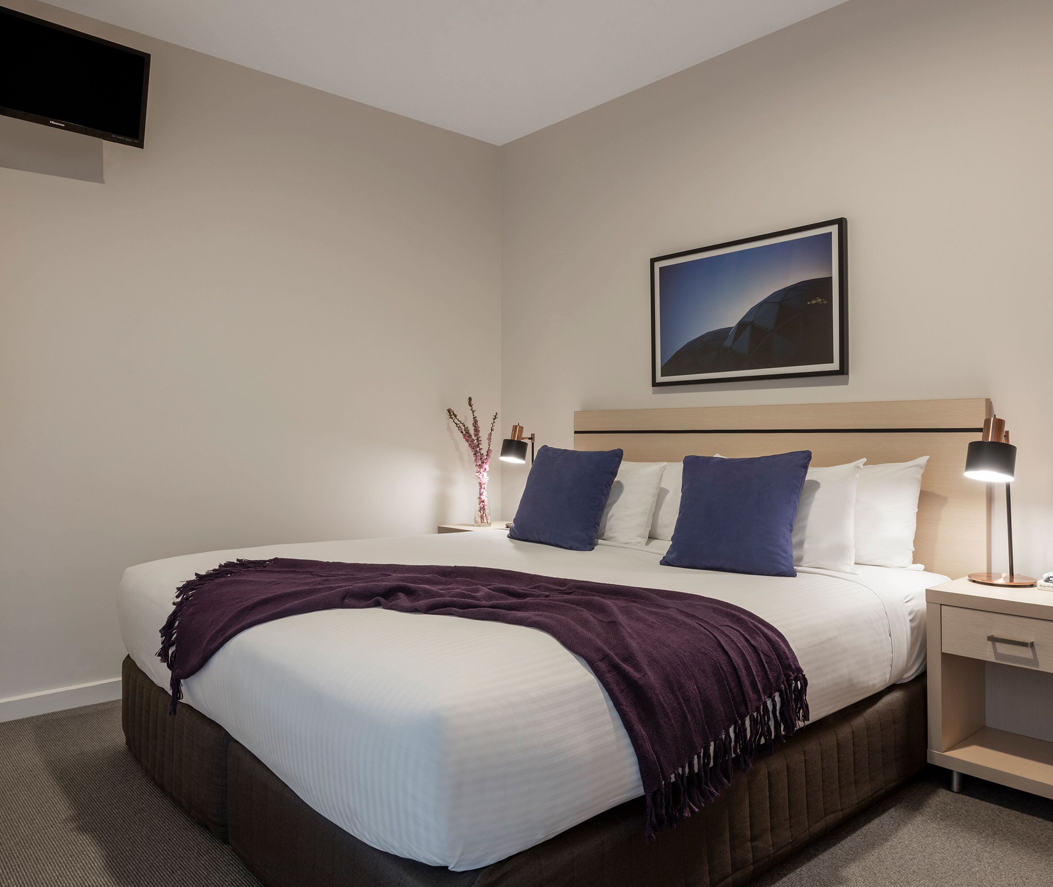 Una habitación de hotel con una cama grande, una mesita de noche con una lámpara, un cuadro en la pared y un florero con flores sobre la mesita.