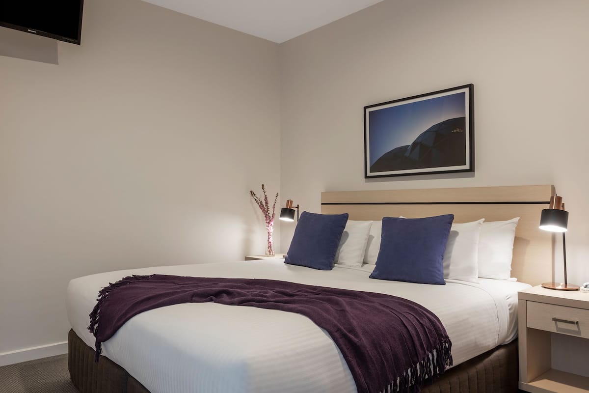 A neatly arranged hotel room with a large bed, a bedside table with a lamp, a framed picture on the wall, and a small vase with flowers.