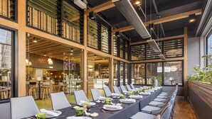 A long dining table set for a meal in a modern restaurant with high ceilings and large windows.
