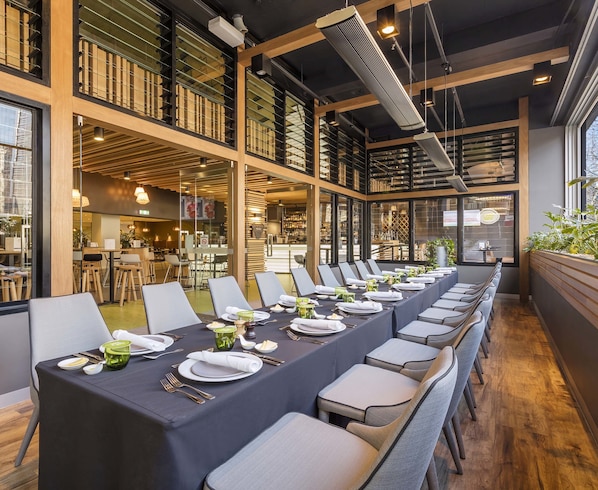 A long dining table set for a meal in a modern restaurant with high ceilings and large windows.