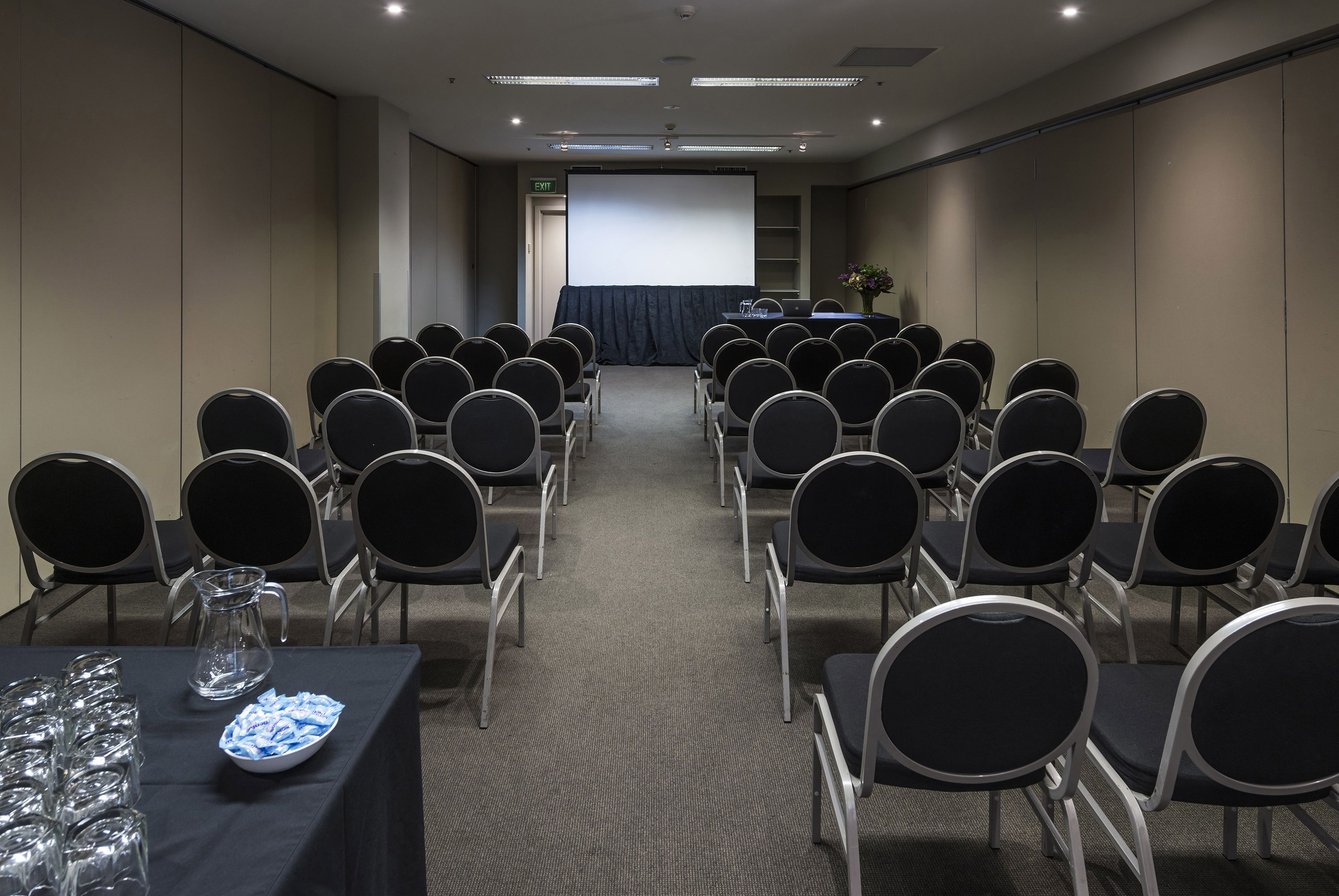 A conference room with rows of chairs facing a projection screen, a table with a pitcher and glasses, and a door marked "EXIT".