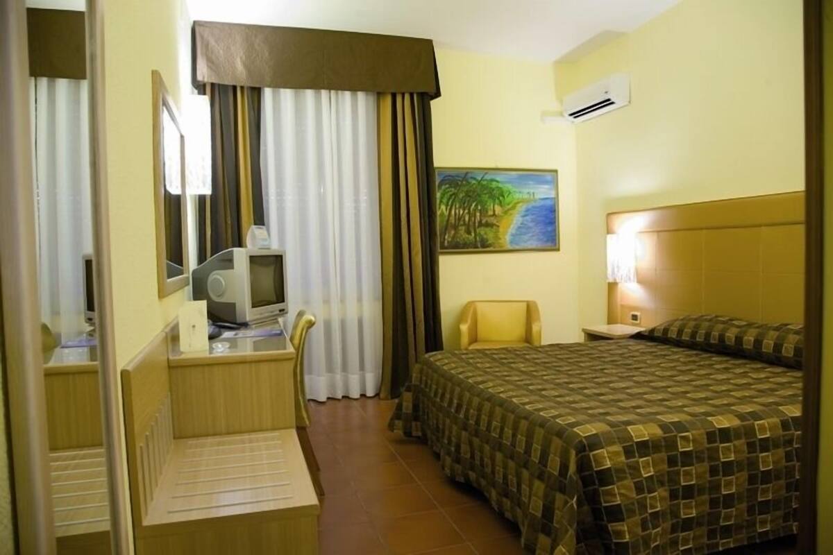 Een hotelkamer met een bed, een bureau met een computer, een stoel, een schilderij van een strand en een airconditioner.