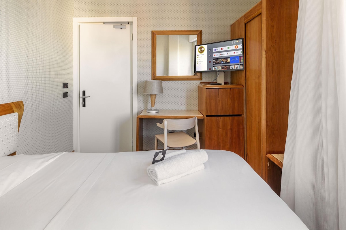 A hotel room with a bed, a desk, a chair, a TV, and a wardrobe.