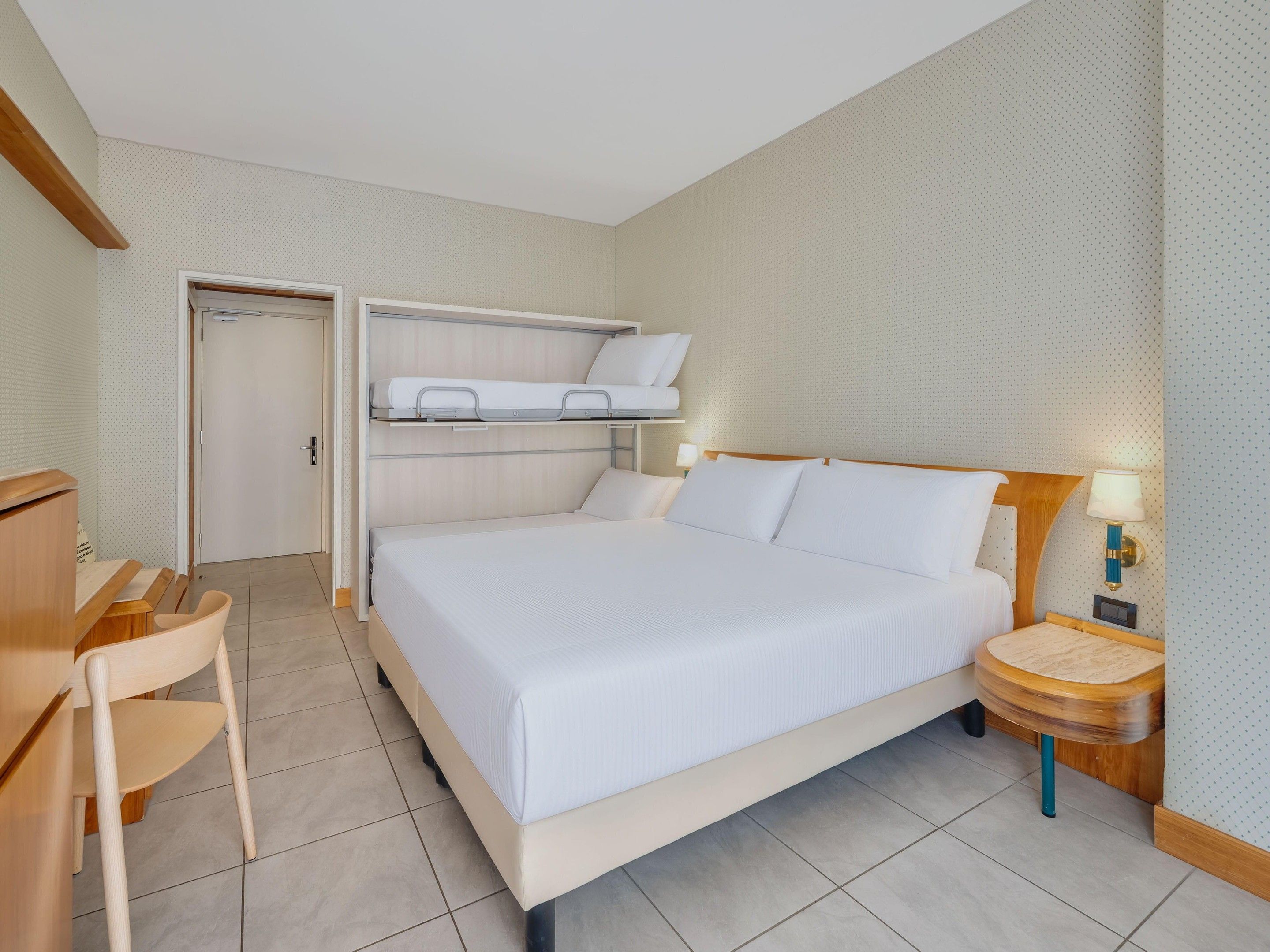 A hotel room with a bunk bed, a desk, a chair, a bedside table with a lamp, and a tiled floor.