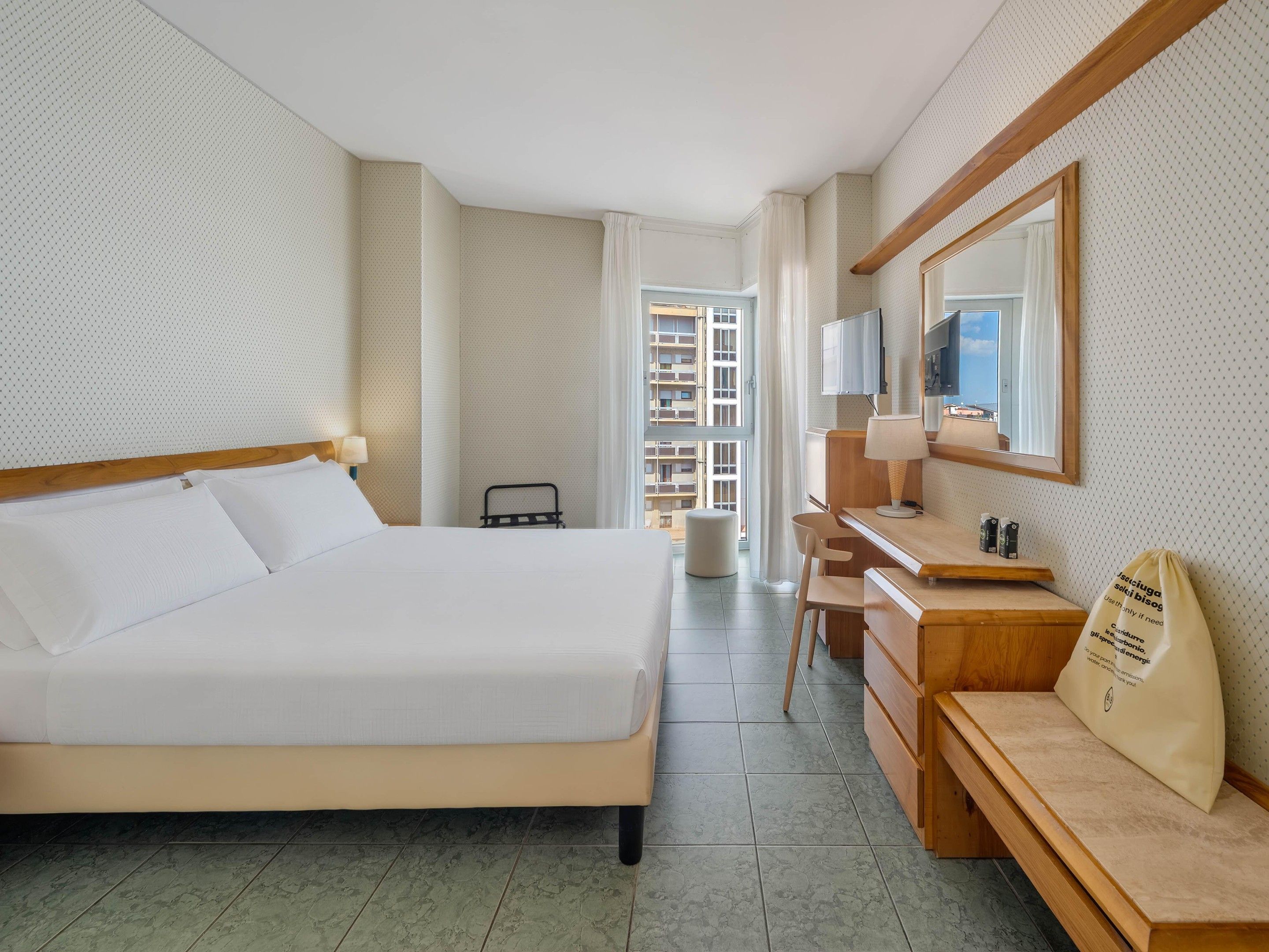 A hotel room with a bed, a desk, a chair, a mirror, and a view of the outside.