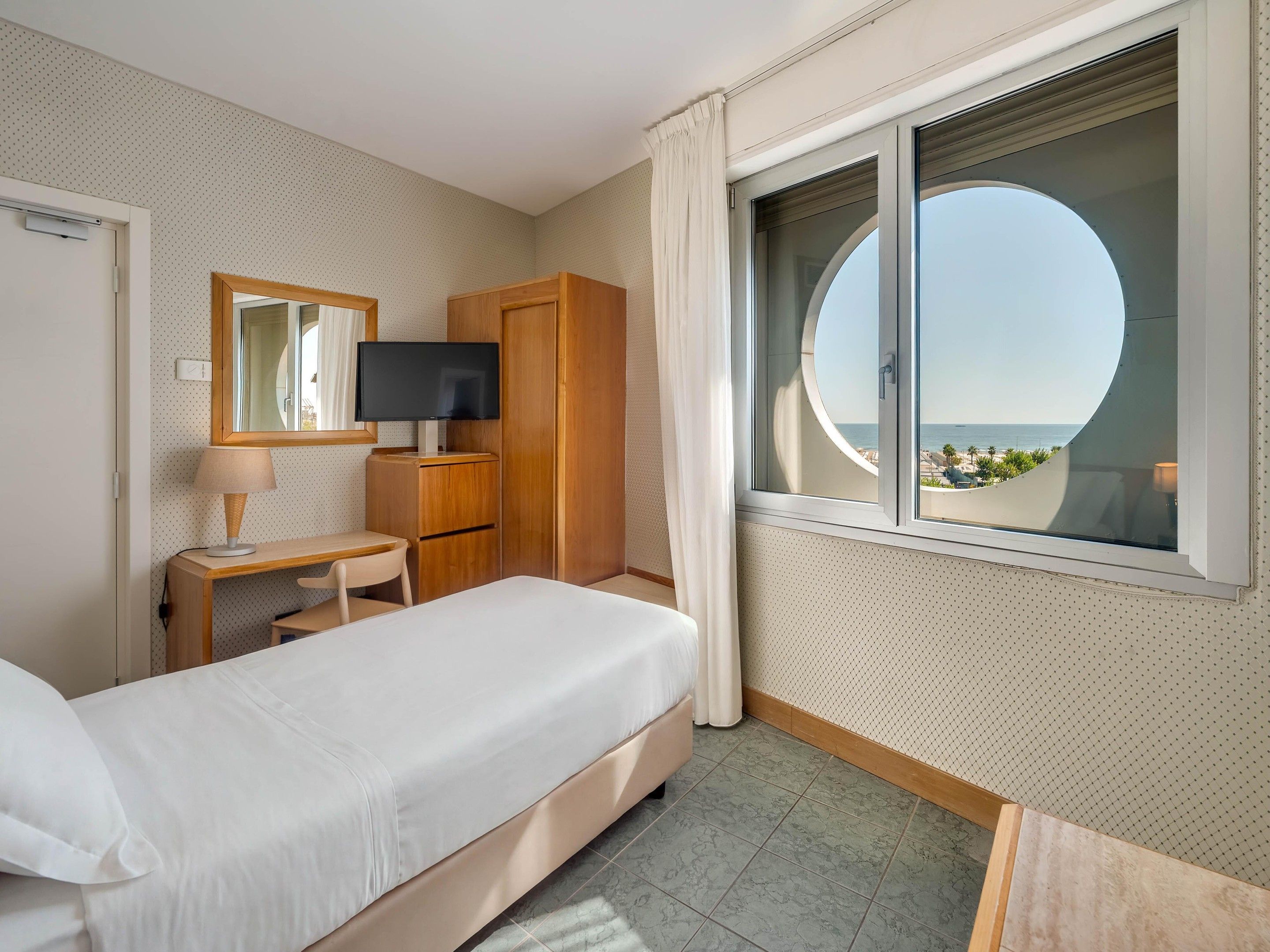 A hotel room with a bed, a desk with a lamp, a TV, a wardrobe, and a window with a view of the beach.