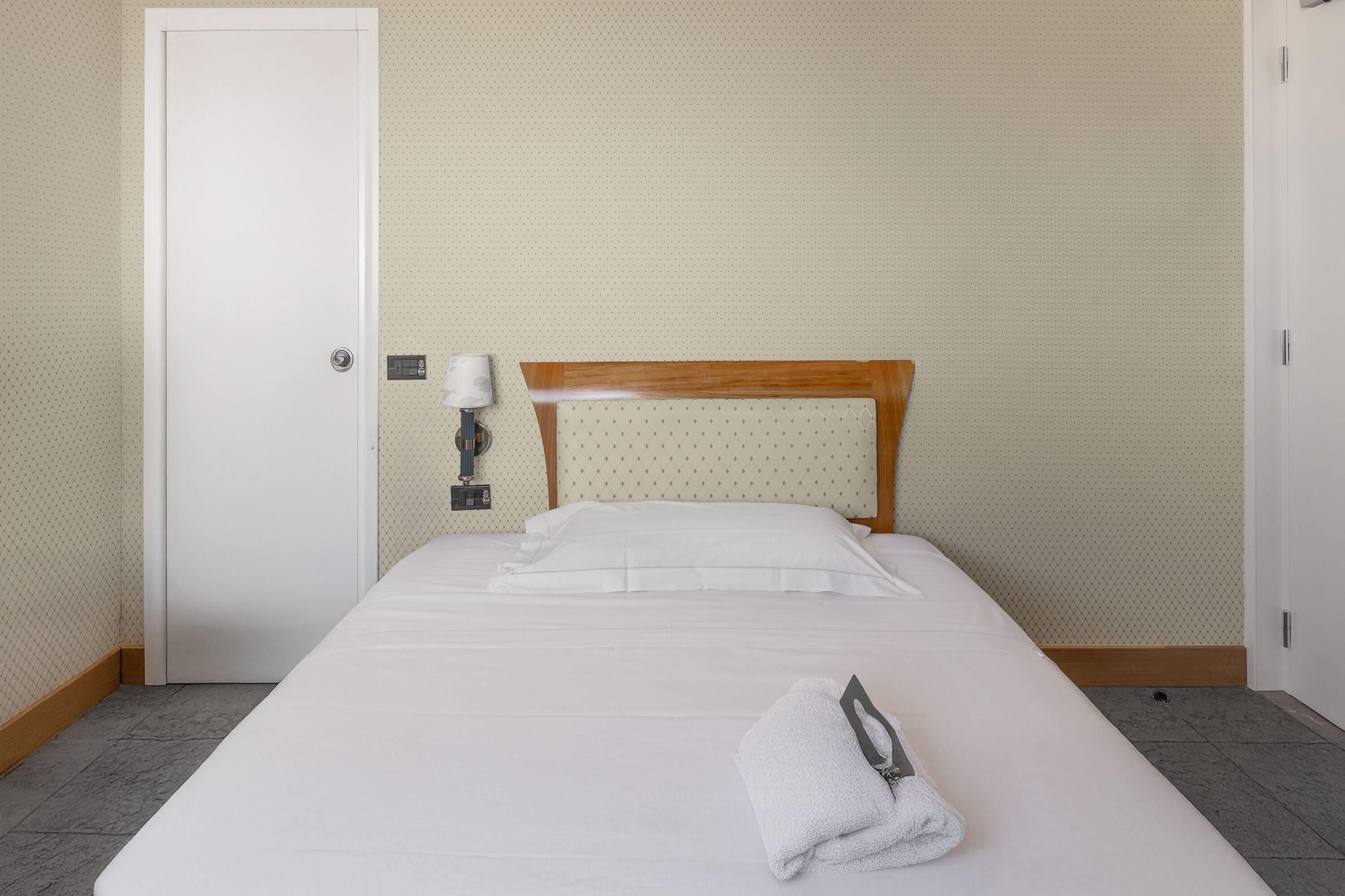 Una cama bien tendida con sábanas blancas y una toalla doblada al lado, cabecera de madera y una puerta blanca con un pequeño panel de control.