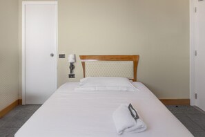 Una cama bien tendida con sábanas blancas y una toalla doblada al lado, cabecera de madera y una puerta blanca con un pequeño panel de control.