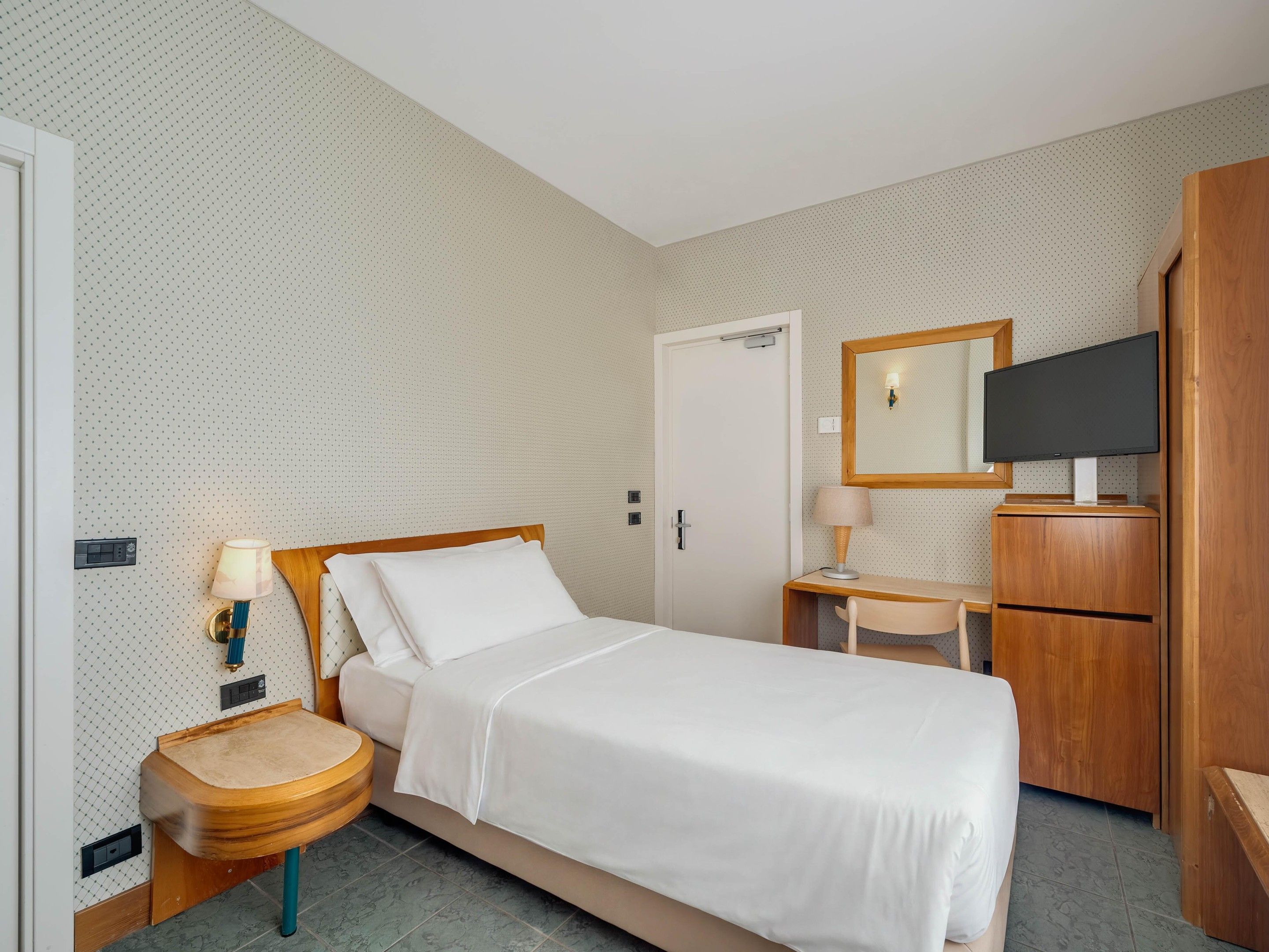 A hotel room with a bed, bedside table, lamp, mirror, TV, and wardrobe.