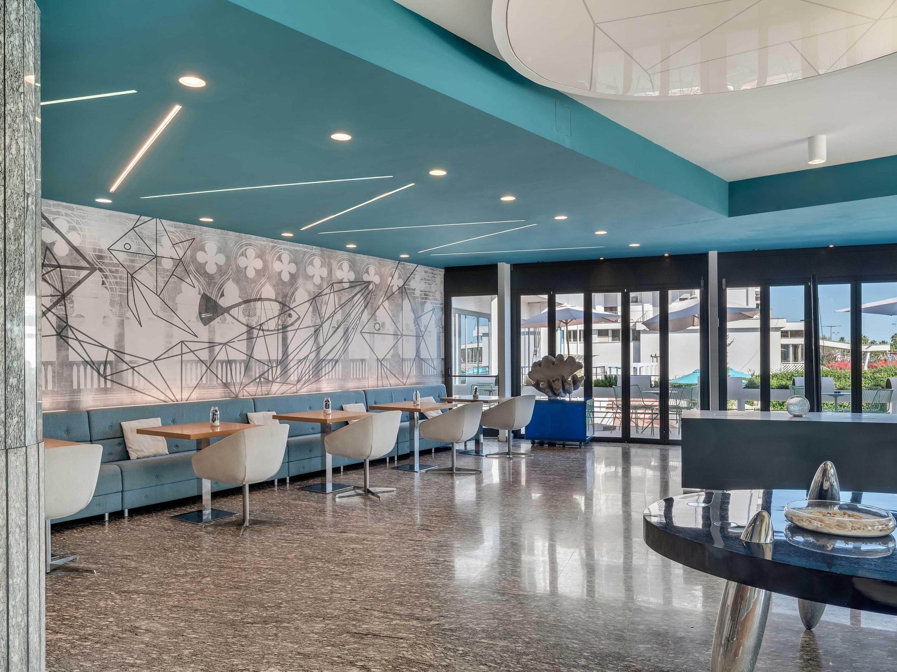 A modern interior with a seating area, a large wall mural, and a view through large windows.