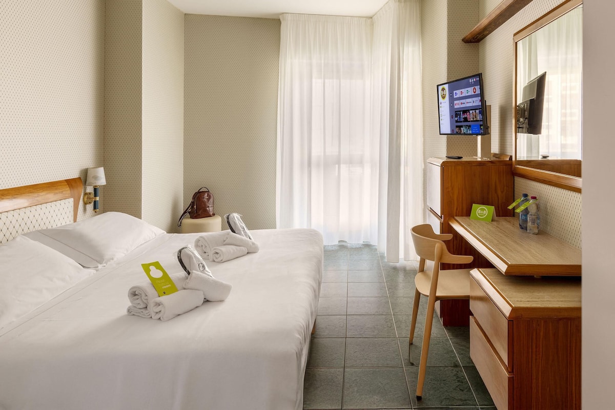 A hotel room with a bed, a desk, a chair, a TV, and a window with curtains.