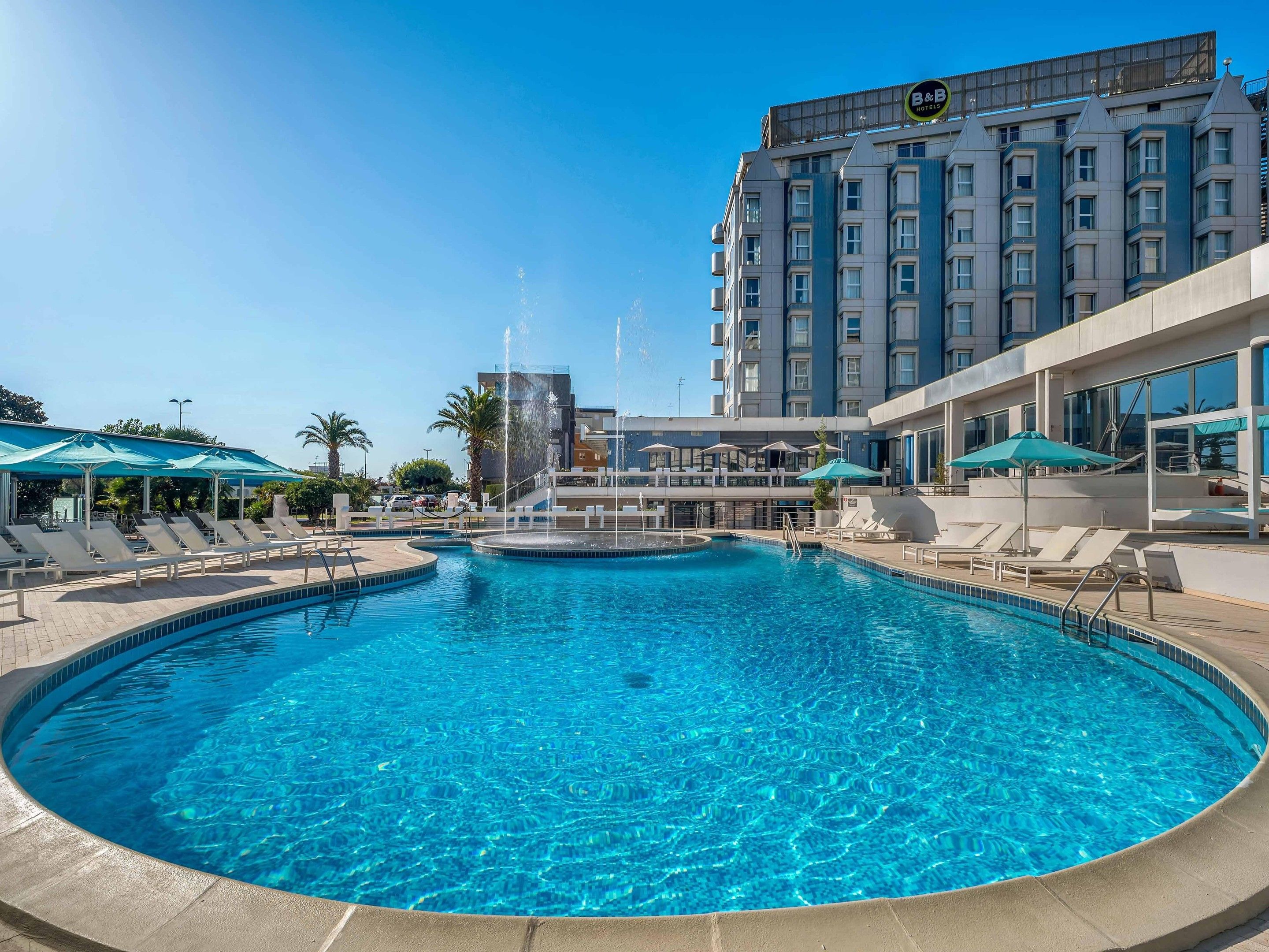 Un espace piscine d'hôtel avec des chaises longues, une fontaine et un bâtiment portant une enseigne.
