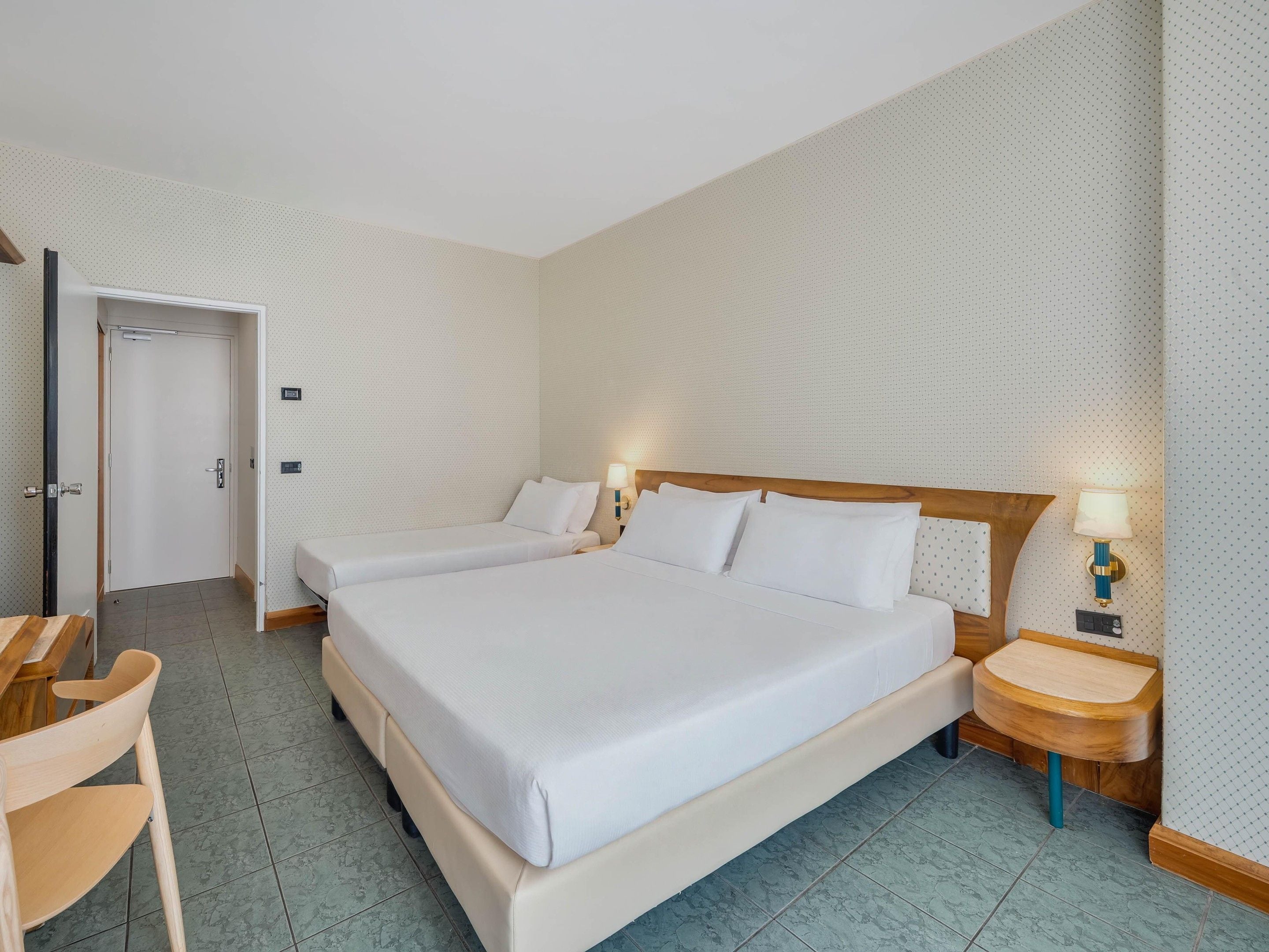 Una habitación de hotel con una cama grande, dos mesitas de noche con lámparas, una silla de madera y una puerta.