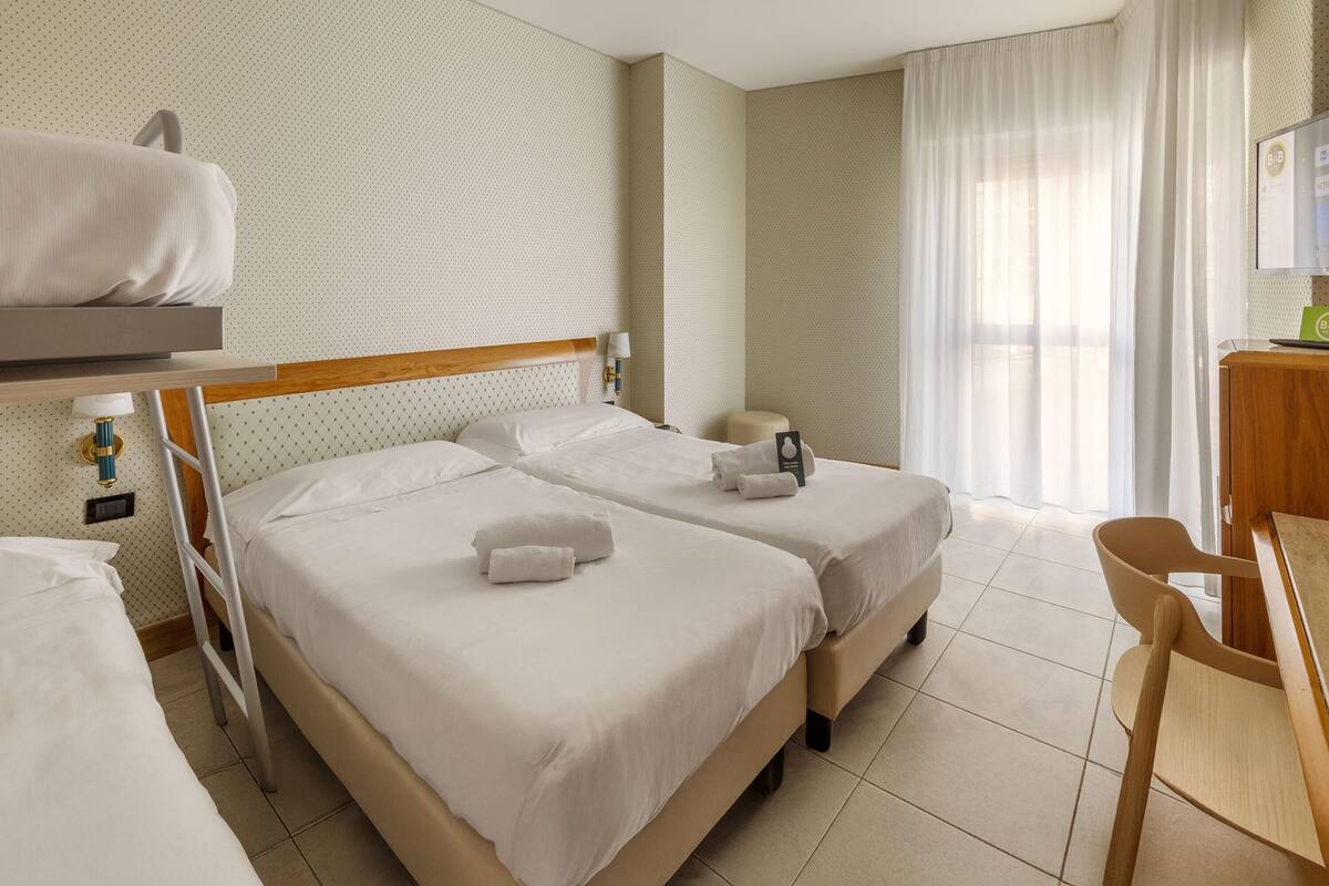 A hotel room with two beds, a desk, a chair, a lamp, and a window with curtains.