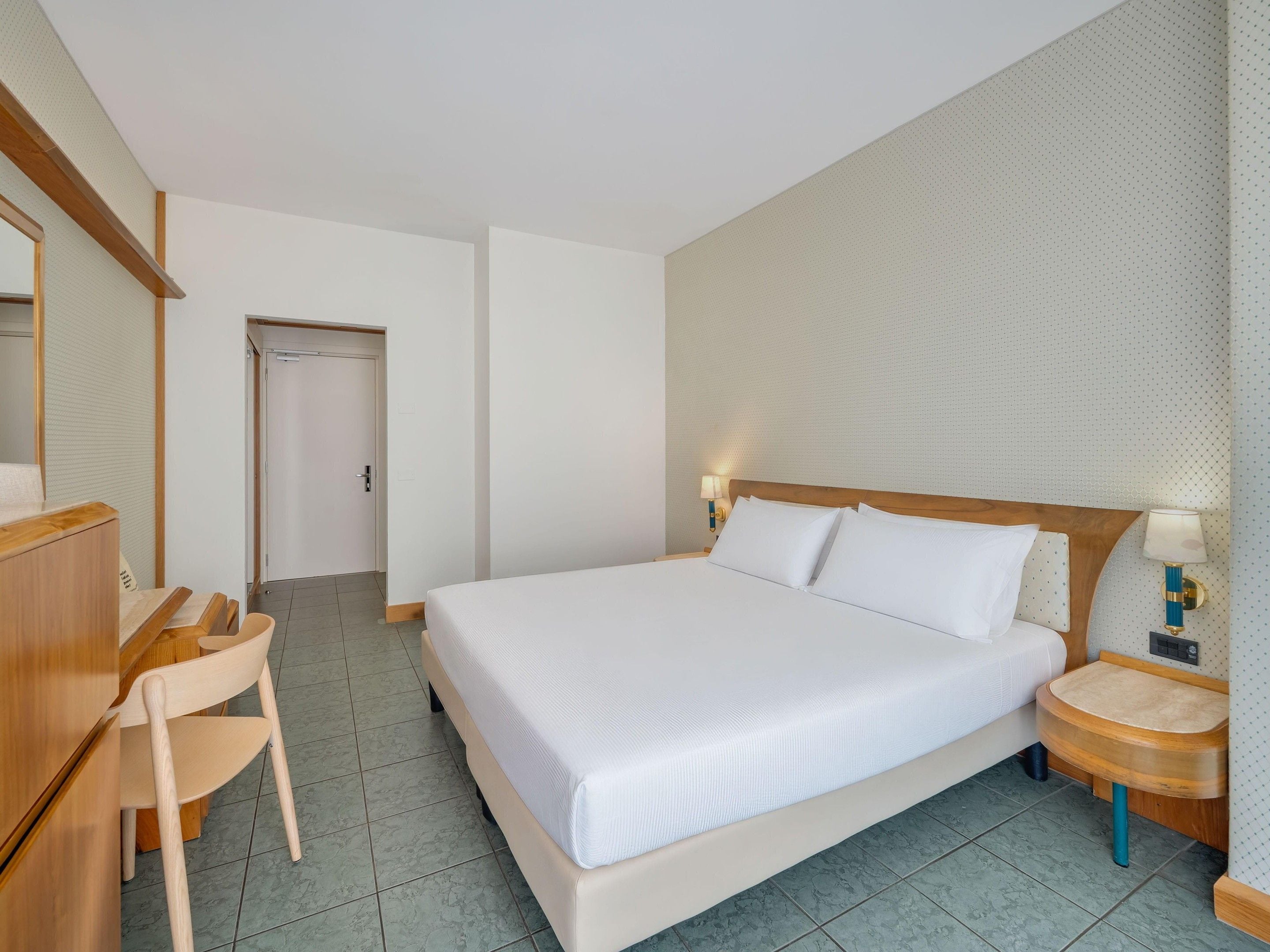 A hotel room with a bed, a desk, a chair, a nightstand, and a wall-mounted lamp.