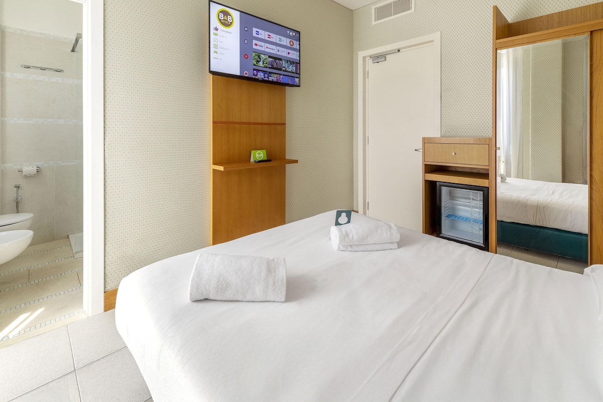 A hotel room with a bed, a TV mounted on the wall, a small shelf, and a mirror.