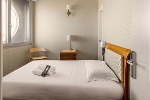 Una cama bien hecha con una toalla doblada y un control remoto, una mesita de noche de madera con una lámpara, una silla y una ventana con persianas.