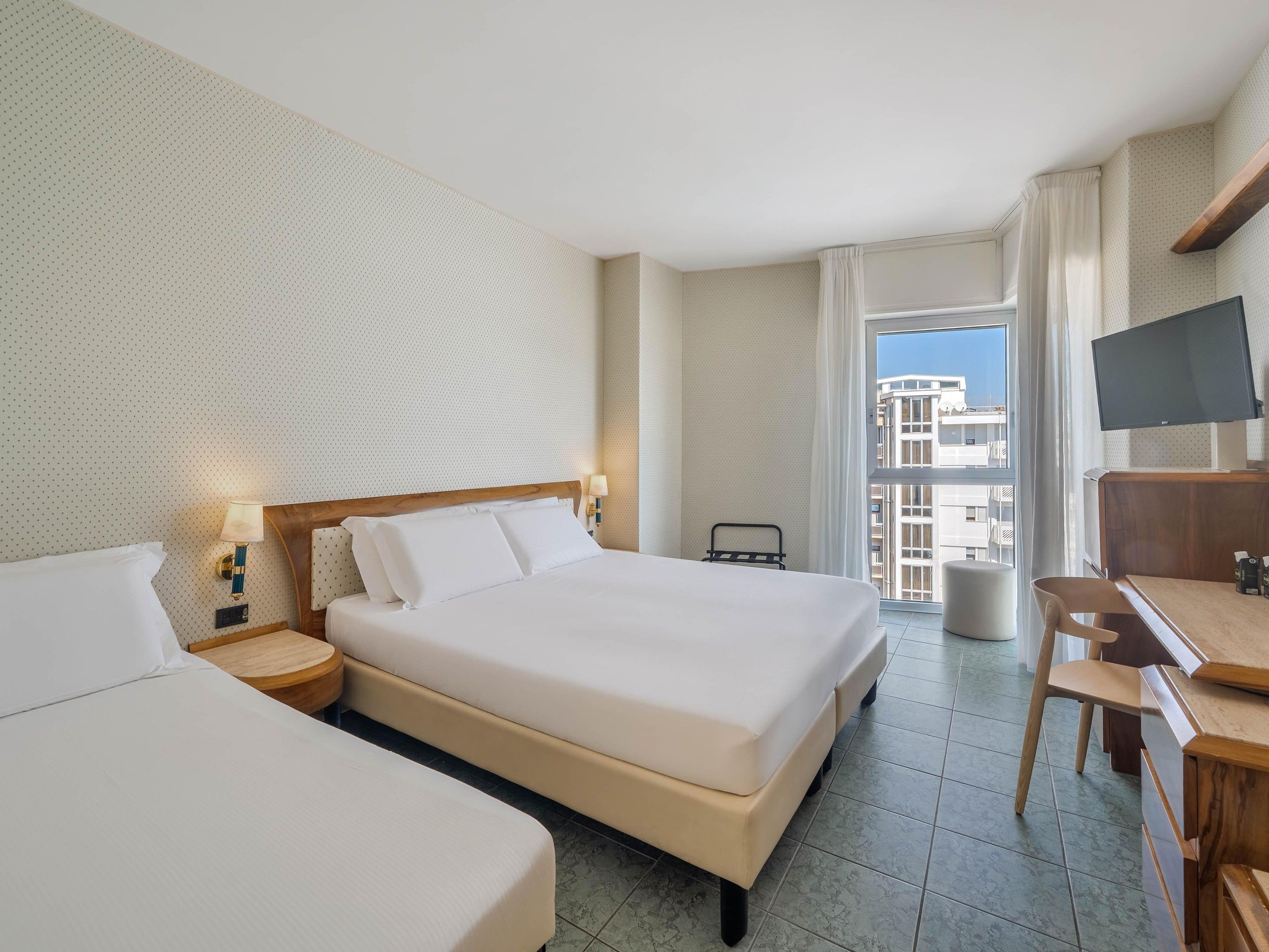 Habitación de hotel con una cama grande, un escritorio, una silla, un televisor y un balcón con vista a la ciudad.