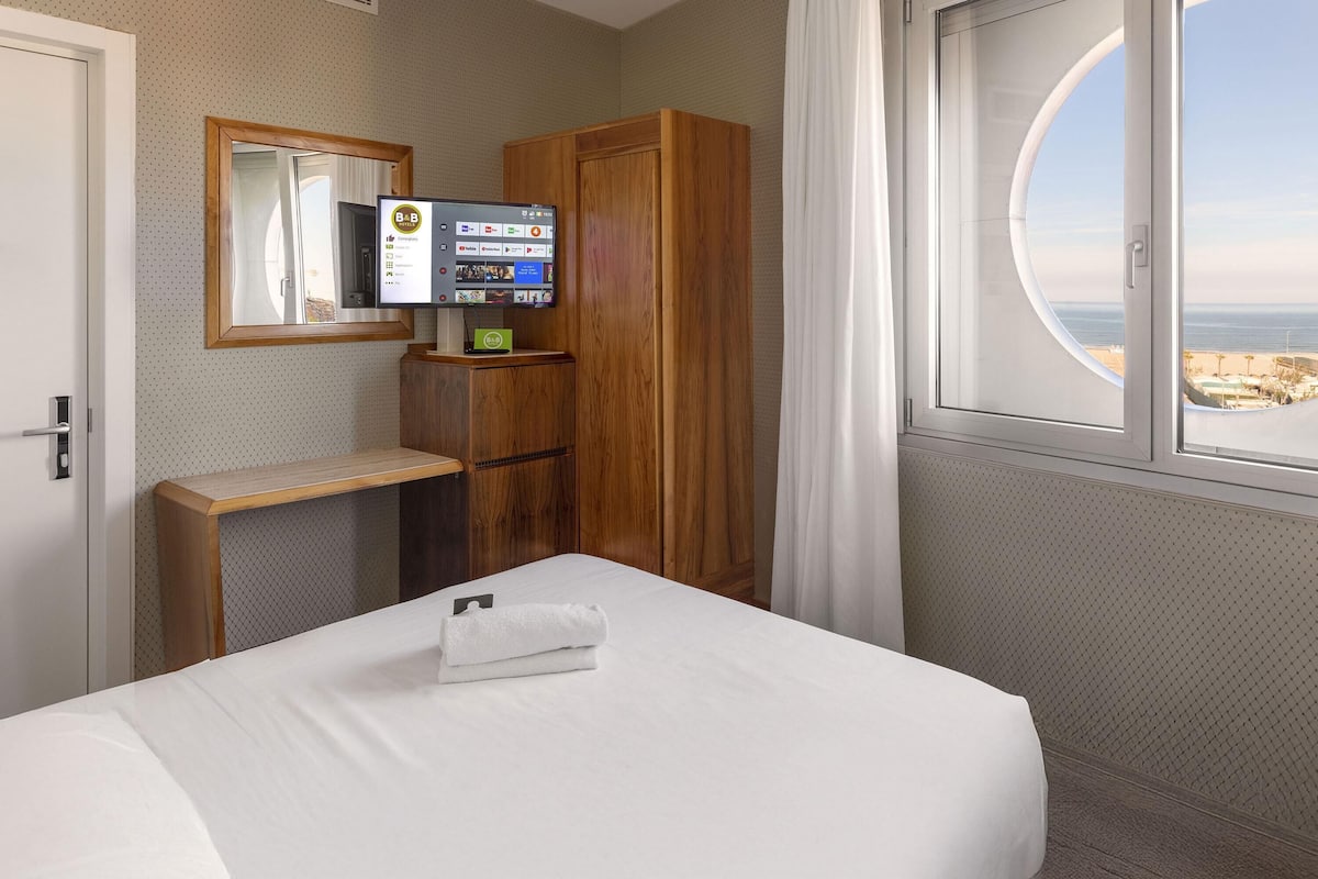 A hotel room with a bed, a wooden wardrobe, a small desk, a television, and a window with a view of the sea.