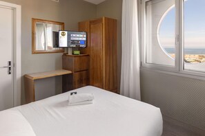 Habitación de hotel con cama, armario de madera, escritorio pequeño, televisor y ventana con vista al mar.