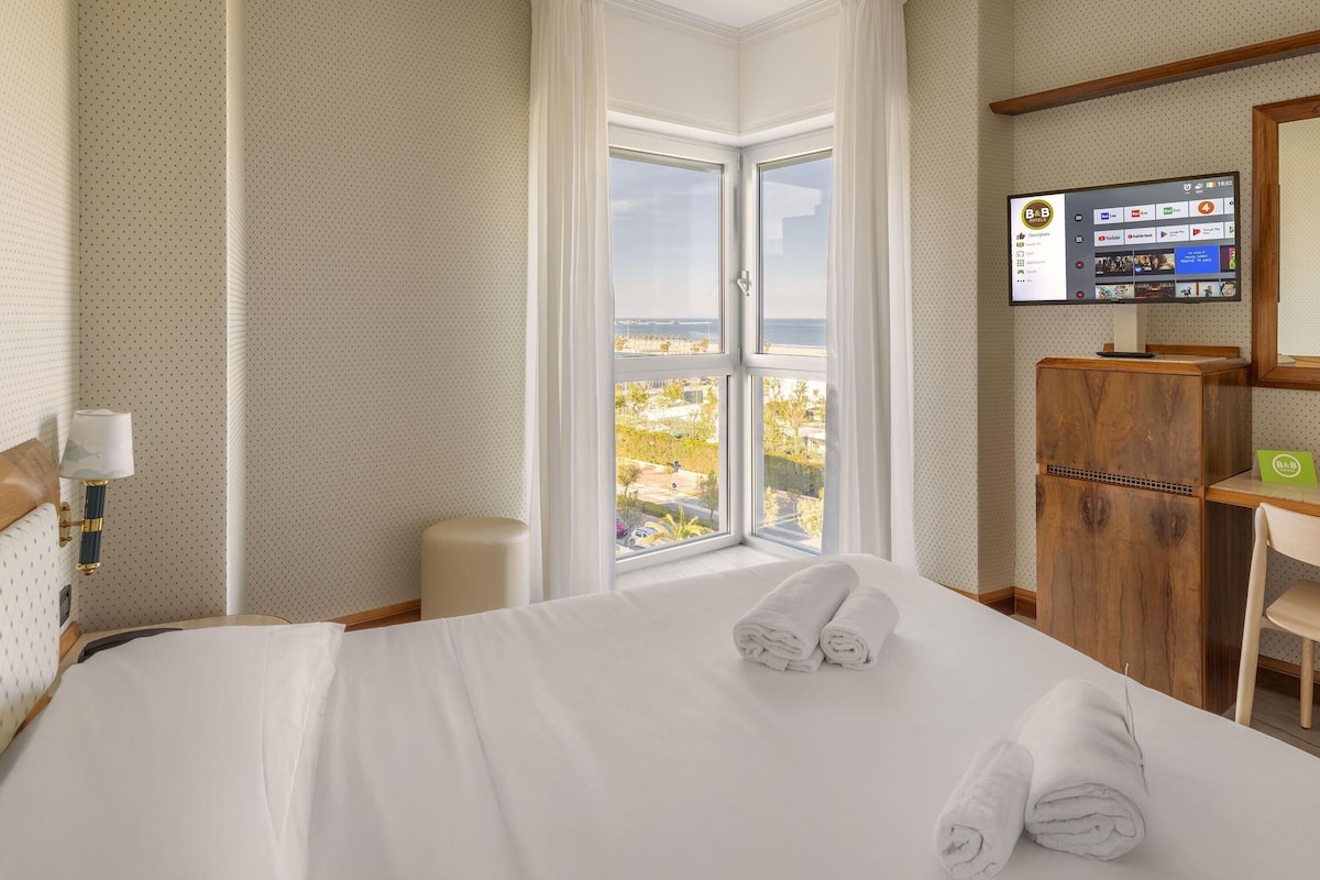 A hotel room with a large bed, a TV mounted on the wall, and a view of the city through a large window.
