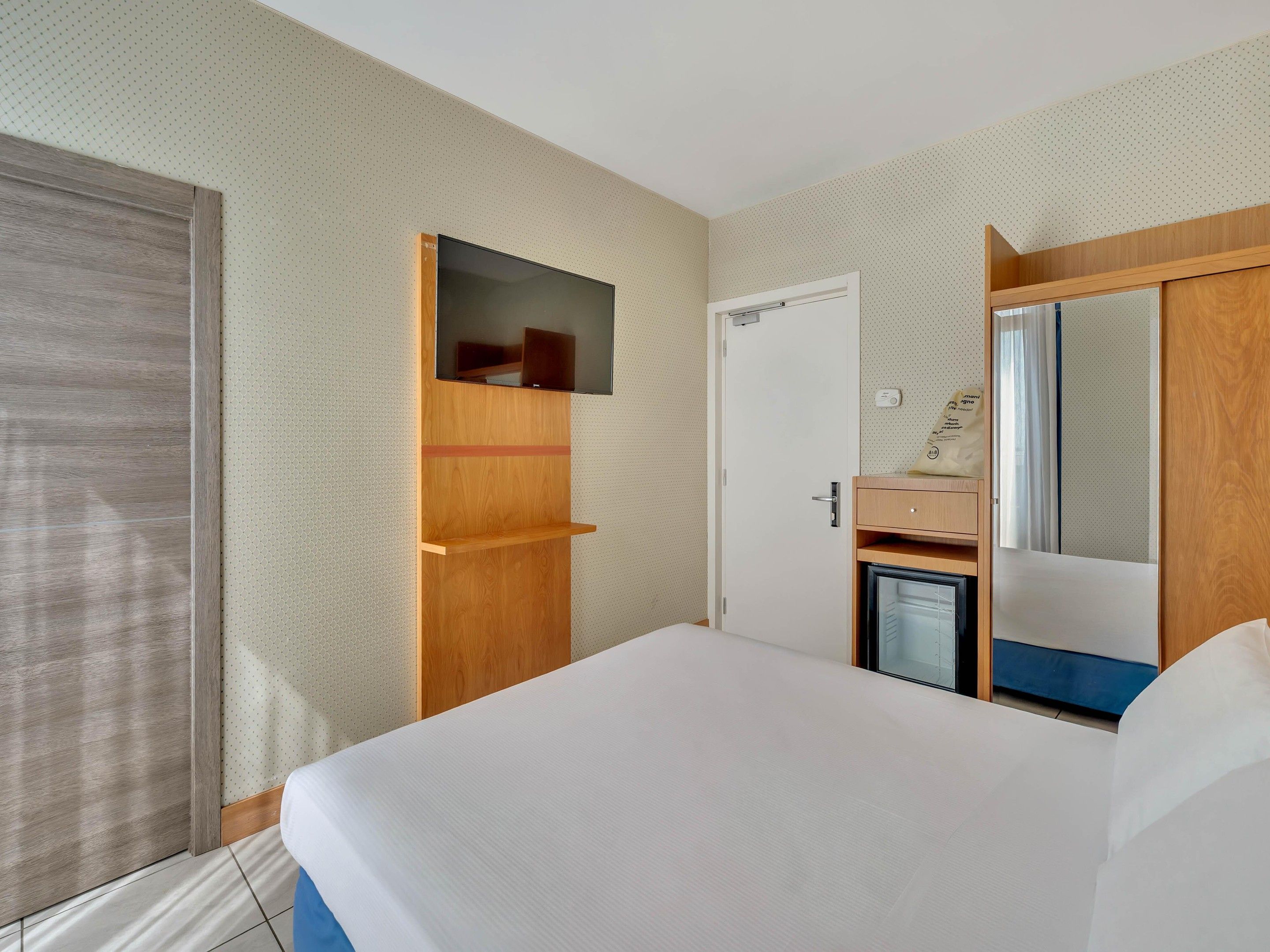 Een hotelkamer met een bed, een aan een houten paneel bevestigde televisie, een deur en een spiegel.