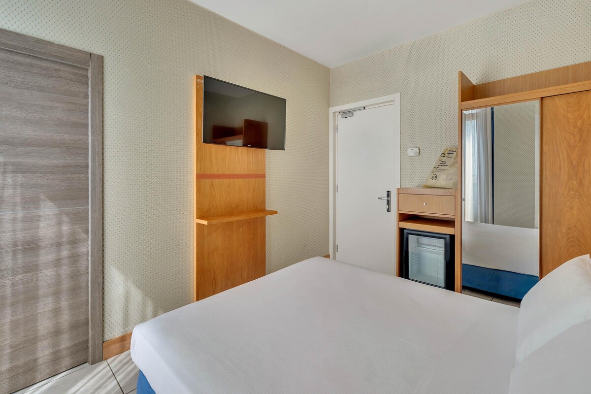 Une chambre d’hôtel avec un lit, un téléviseur fixé sur un panneau en bois, une porte et un miroir.