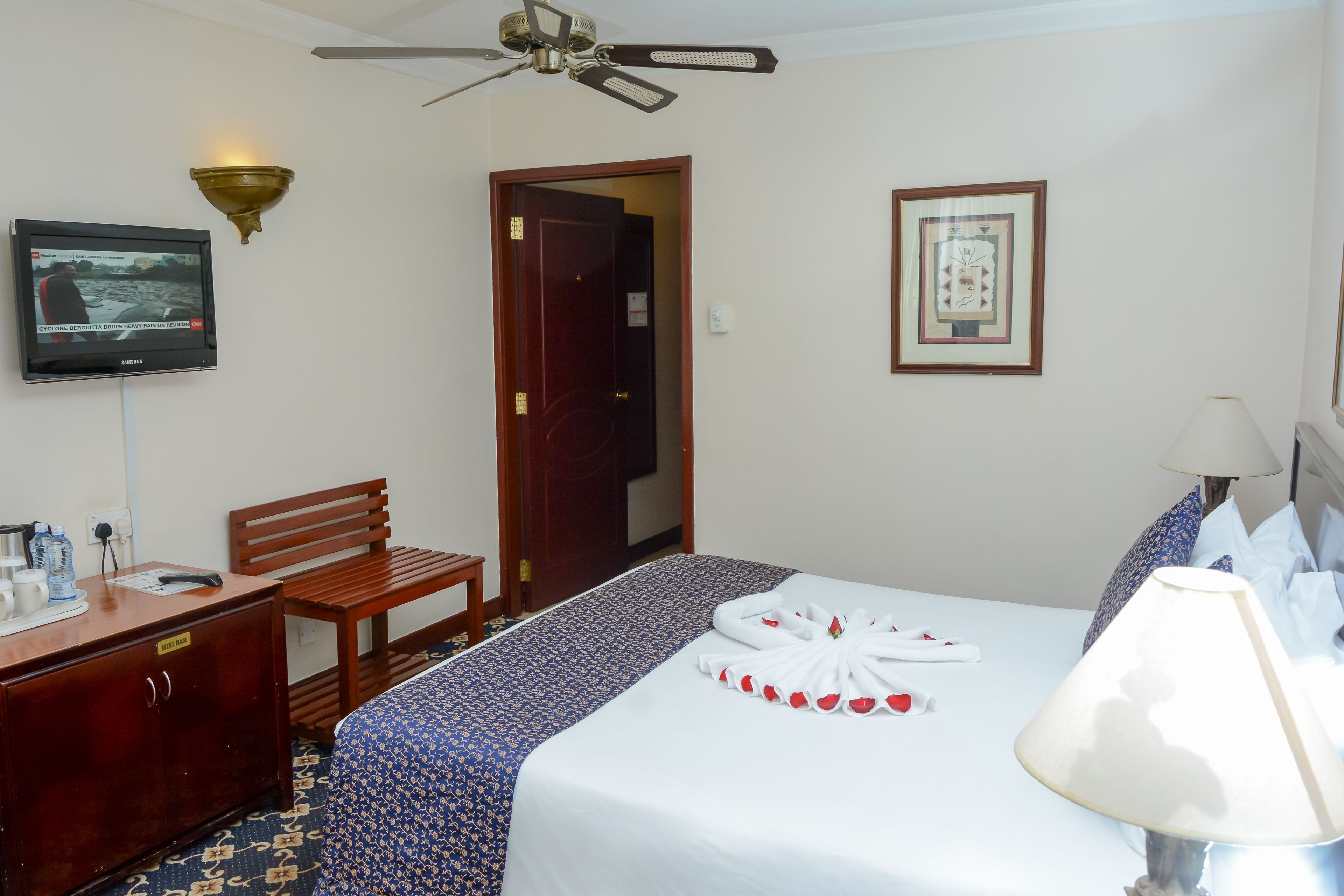 A hotel room with a bed, a desk, a chair, a television, a ceiling fan, and a framed picture on the wall.