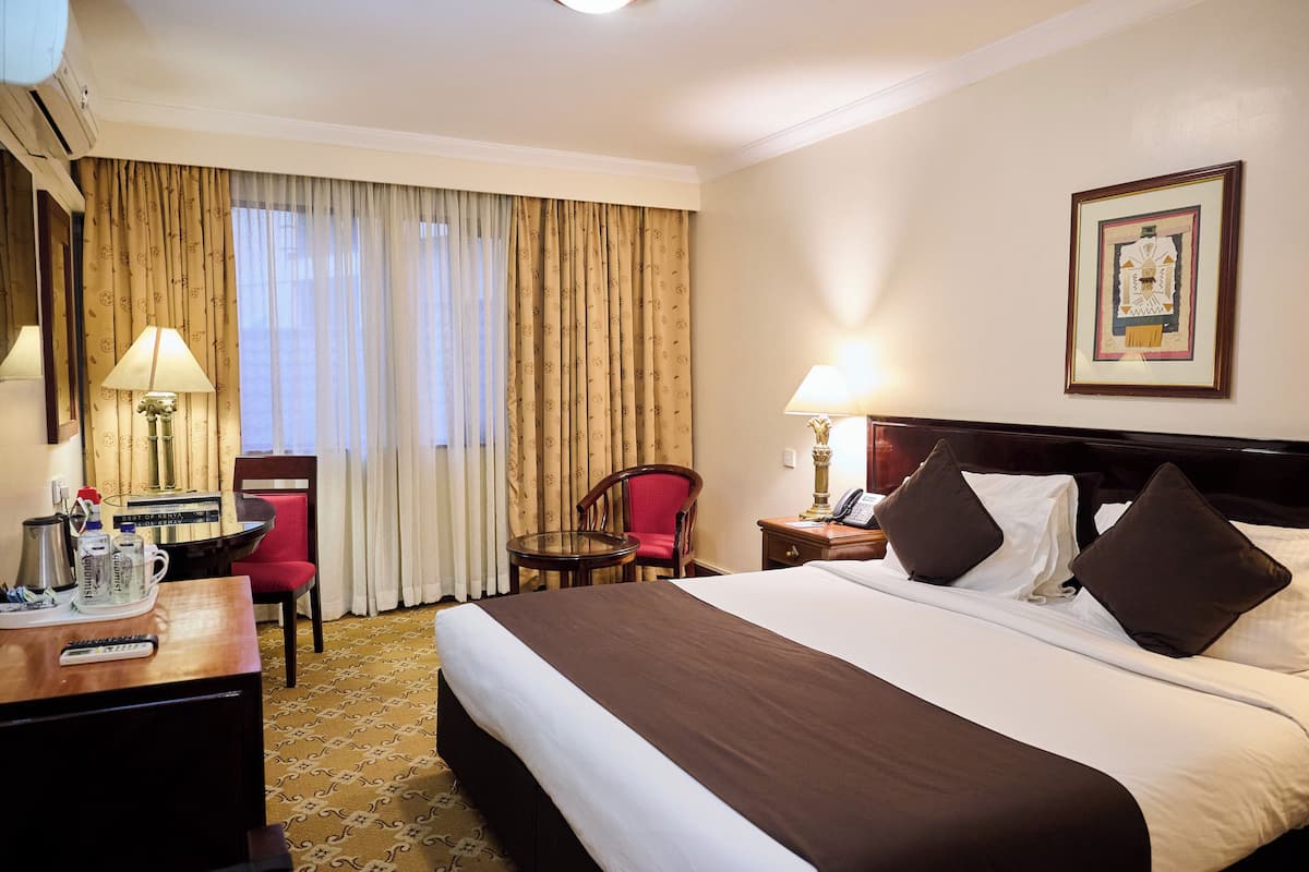 A hotel room with a large bed, a desk with a lamp, a chair, and a small table.