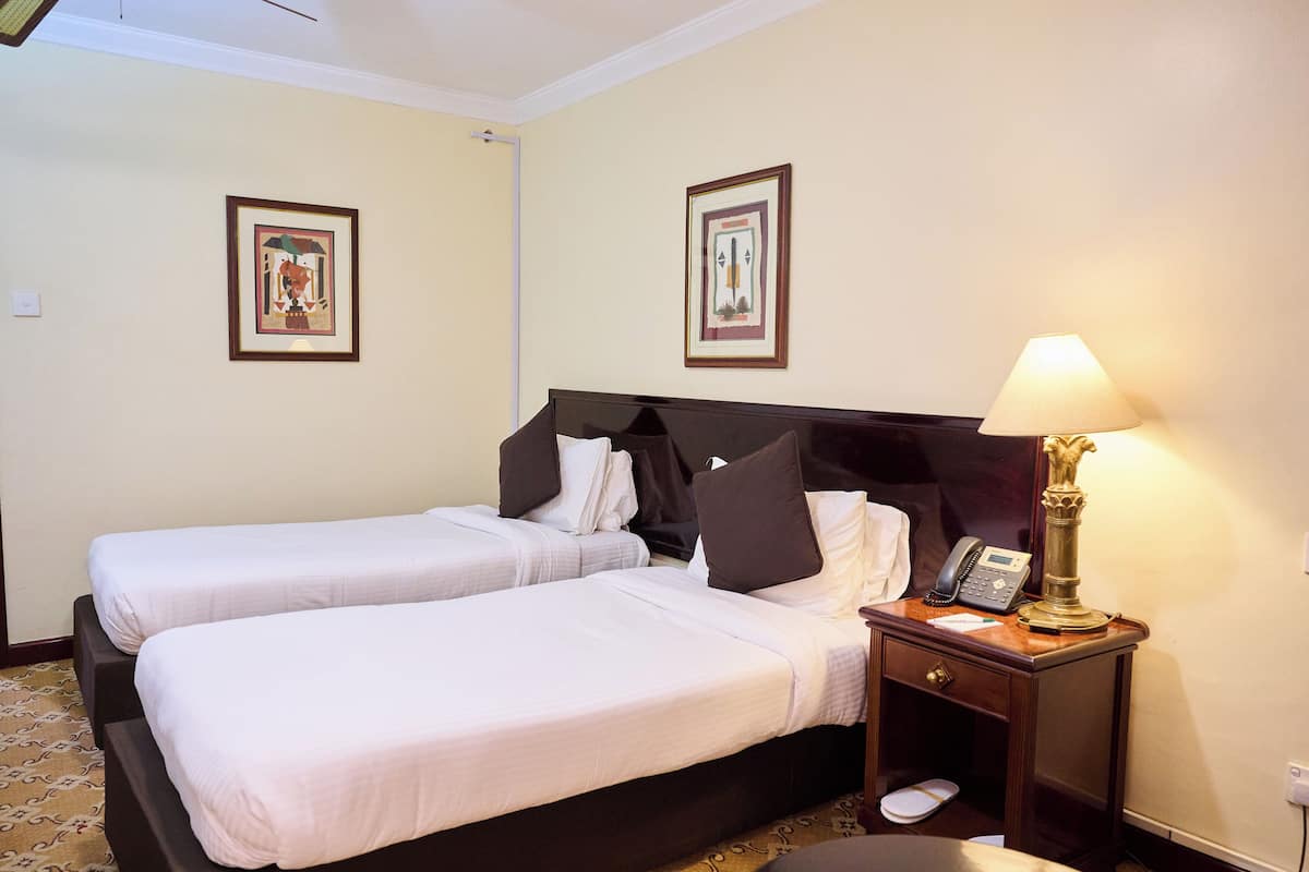 A hotel room with two single beds, a bedside table with a lamp, a telephone, and a small drawer.