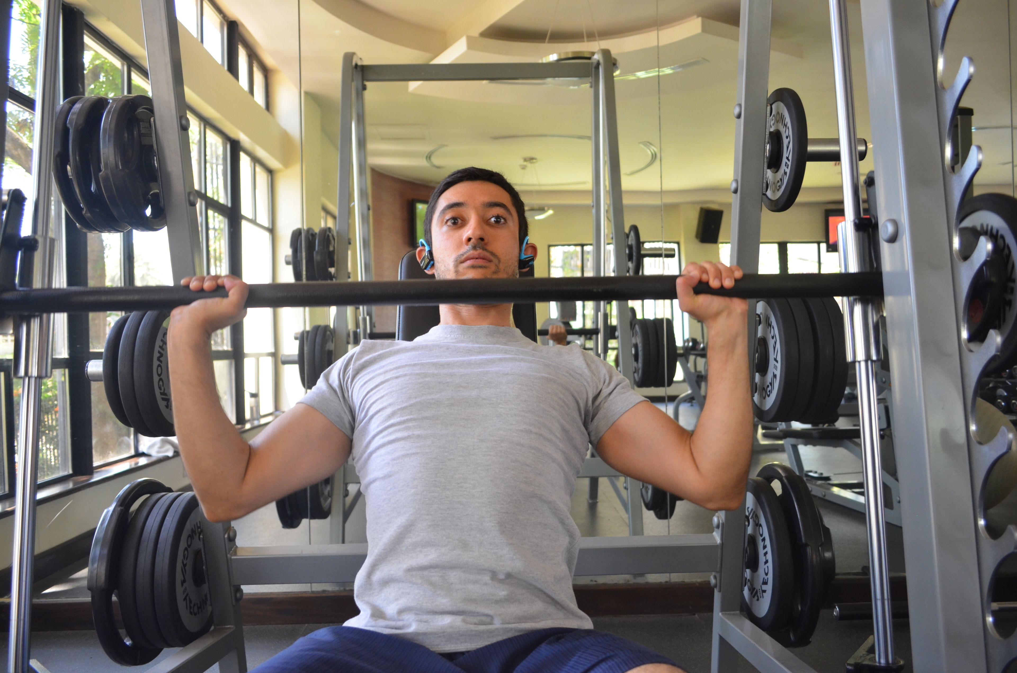 A person performing a bench press exercise in a gym with weightlifting equipment and large windows.