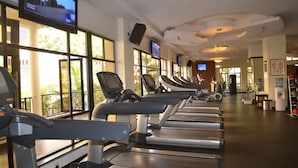 A gym with treadmills lined up along a wall with windows, a television mounted on the wall, and exercise equipment.