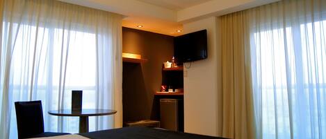 Quarto de hotel com cama, uma pequena mesa, uma cadeira, televisão fixada na parede e área de cozinha compacta.