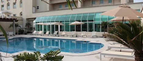 Piscina con sillones de descanso y sombrillas, rodeada por un edificio de fachada de vidrio y palmeras.