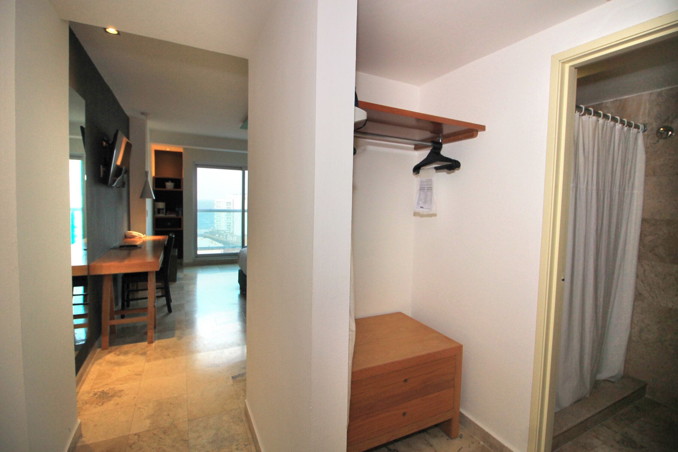 Habitación de hotel con un banco de madera, un mueble pequeño de madera, un perchero con una prenda colgada y una zona de ducha con cortina.