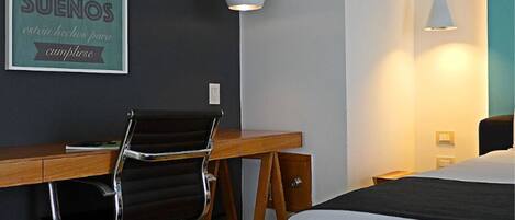 A hotel room with a wooden desk, a black office chair, a bed with white bedding, and a wall-mounted light fixture.