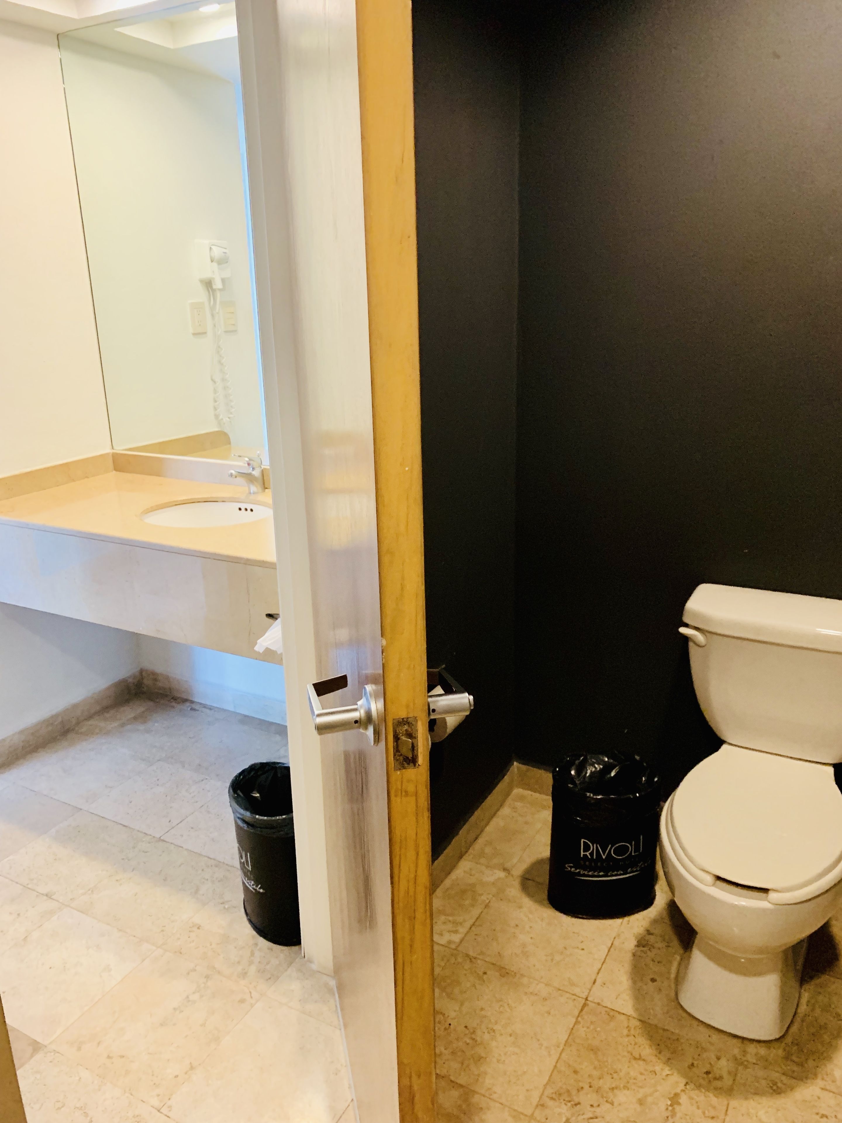 Un baño con inodoro blanco, un bote de basura negro y una botella de limpiador RIVOLI.