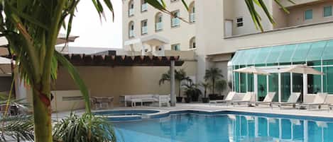 Una piscina con sillas de descanso y sombrillas, rodeada de palmeras y un edificio con varias ventanas.