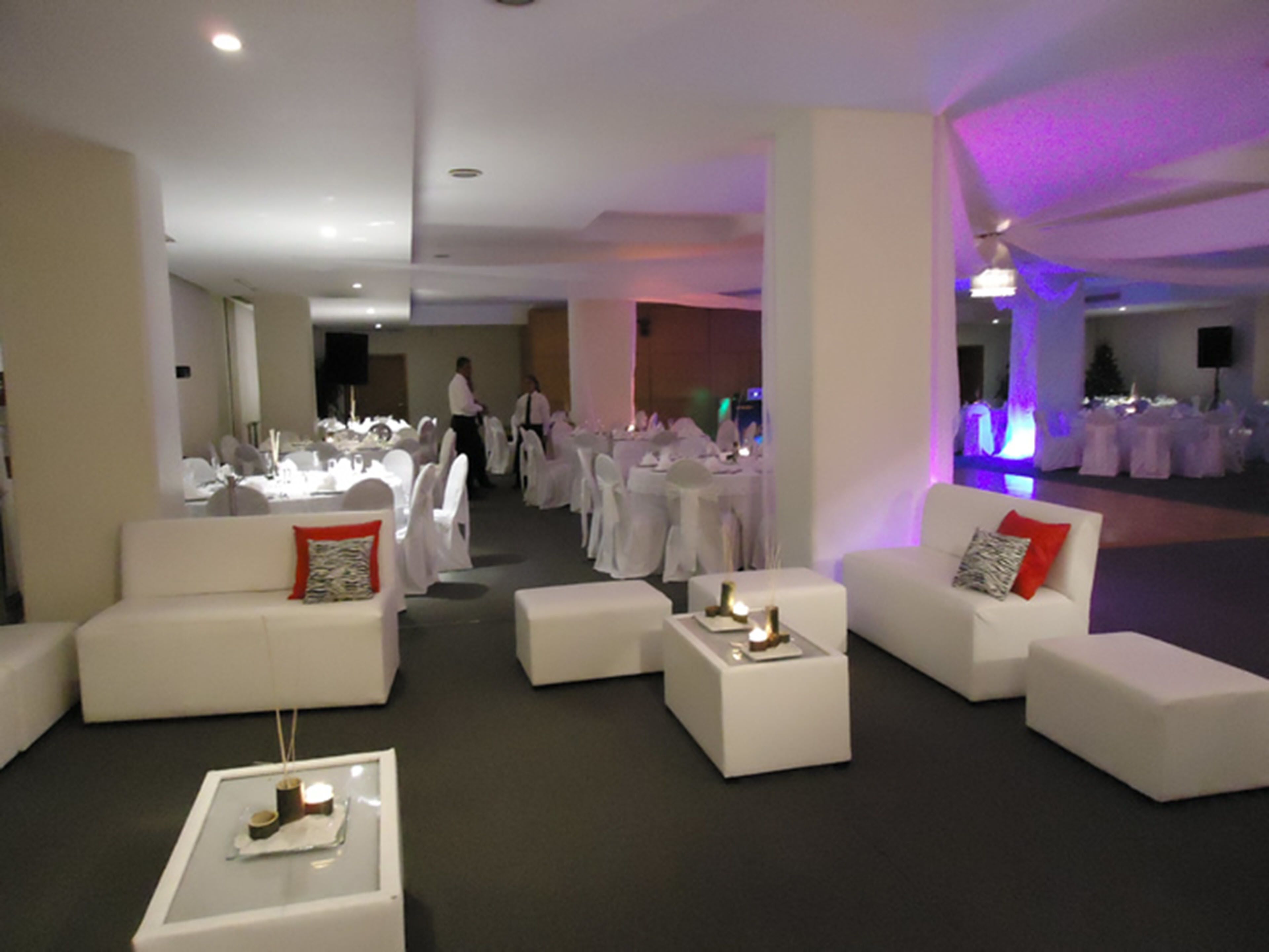 A spacious lounge area with white sofas and tables, a long table with chairs set up for dining, and a stage area in the background.
