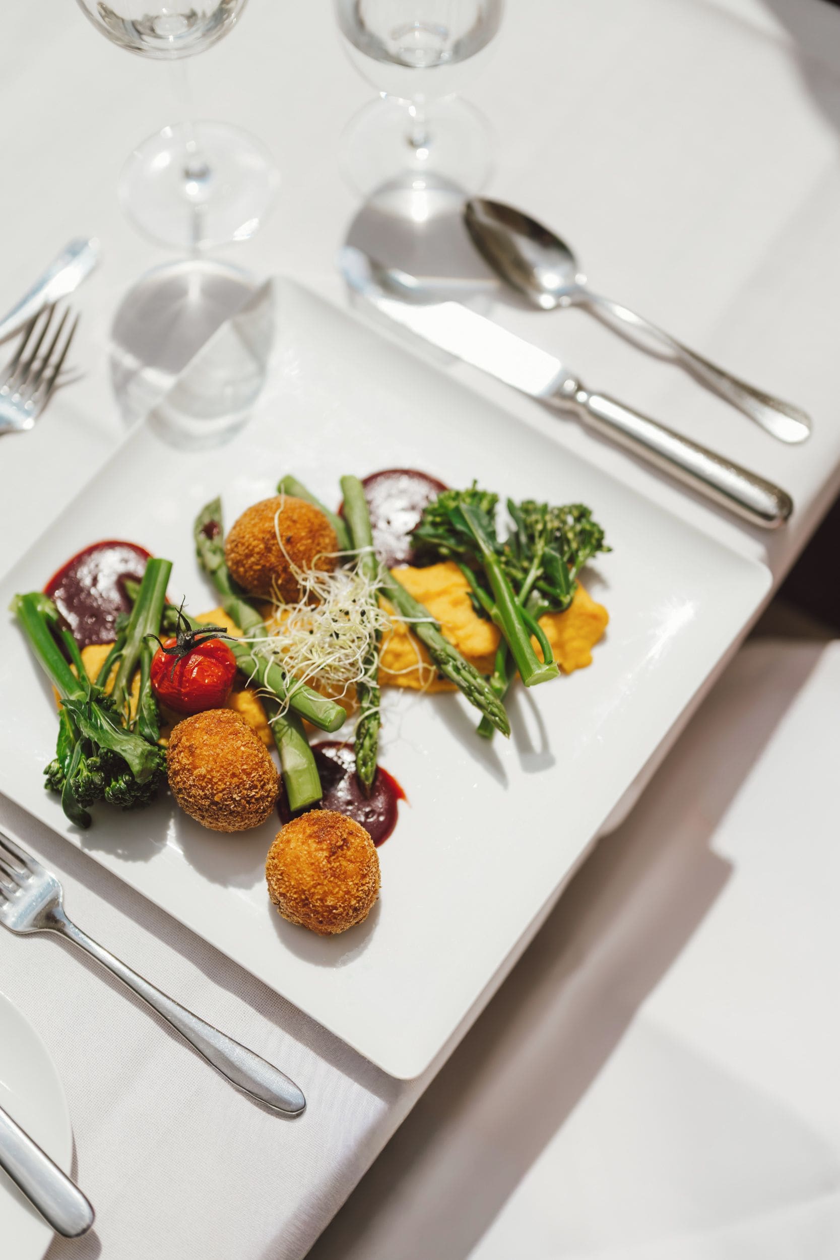 Ein angerichtetes Essen mit verschiedenen Gemüsesorten und einem panierten Gericht, begleitet von Besteck und Gläsern auf einem Tisch.