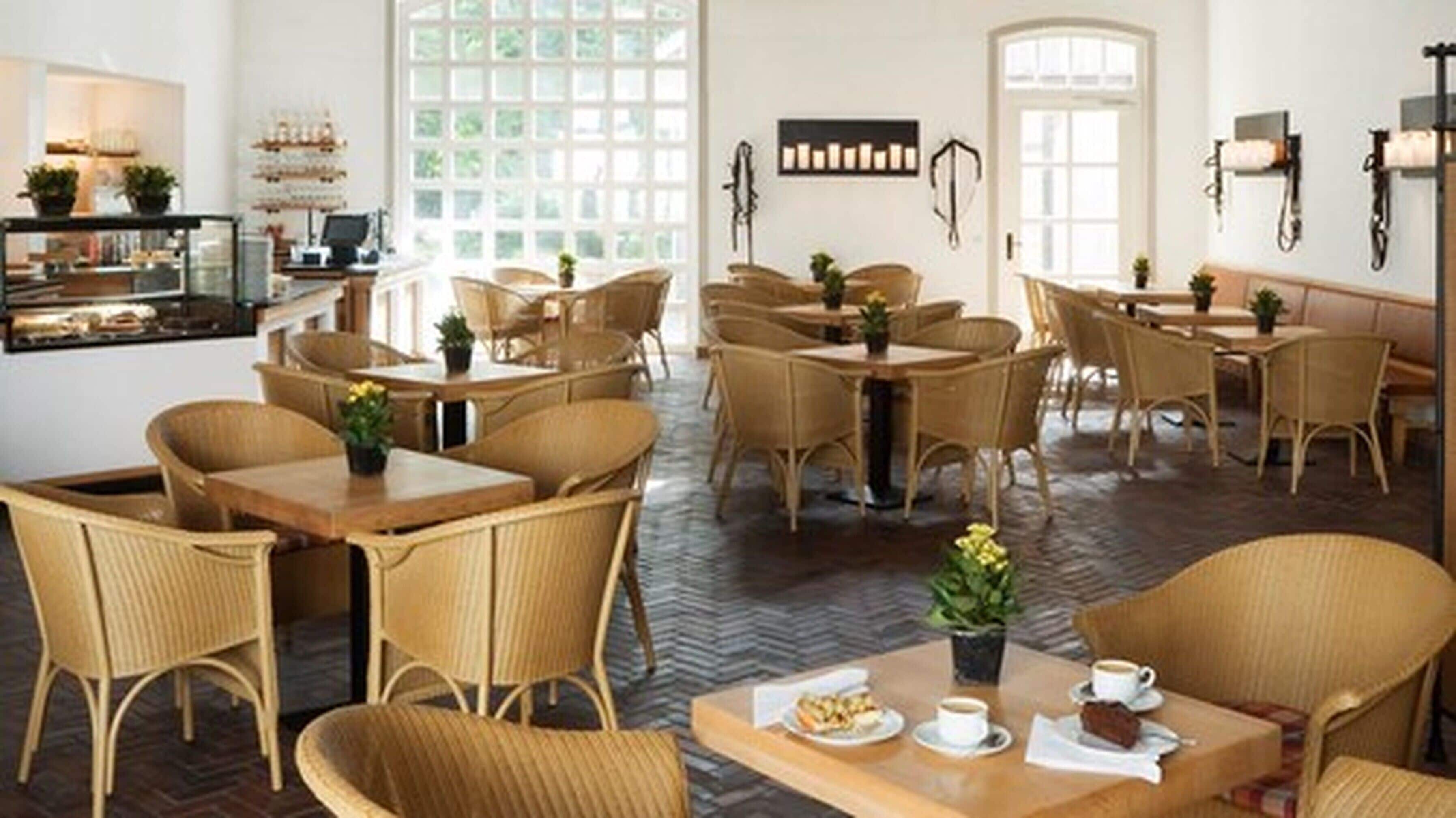 Un café amplio con mobiliario de mimbre, ventanas grandes y una zona de cocina a la vista.