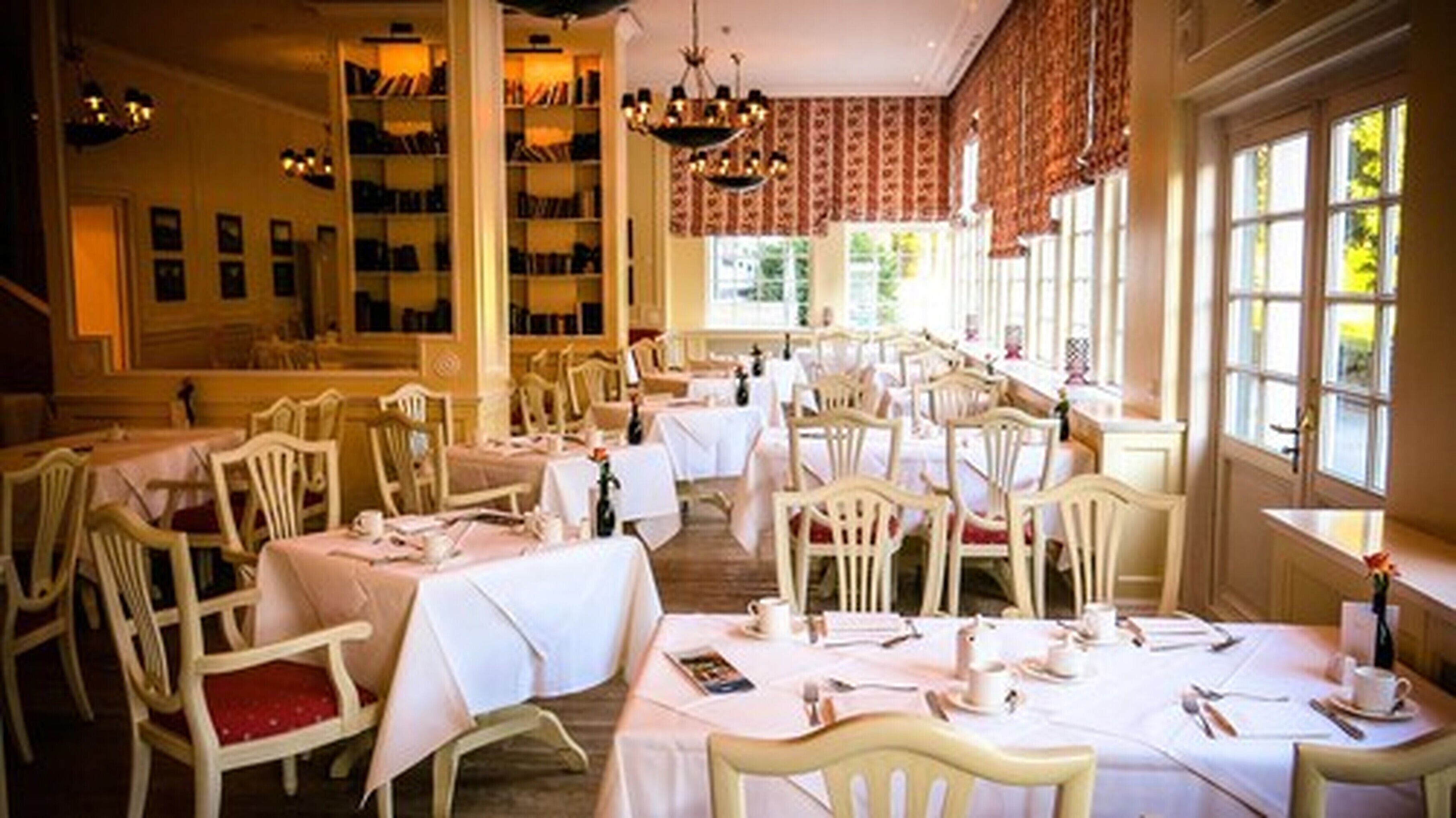 A dining area with multiple tables and chairs, a chandelier, and large windows with curtains.