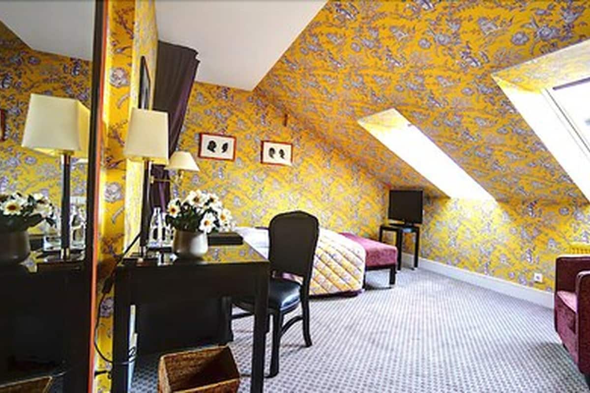 Ein Zimmer mit schrägen Wänden, einem Bett, einem Schreibtisch mit Stuhl und einem kleinen Tisch mit Blumenvase.