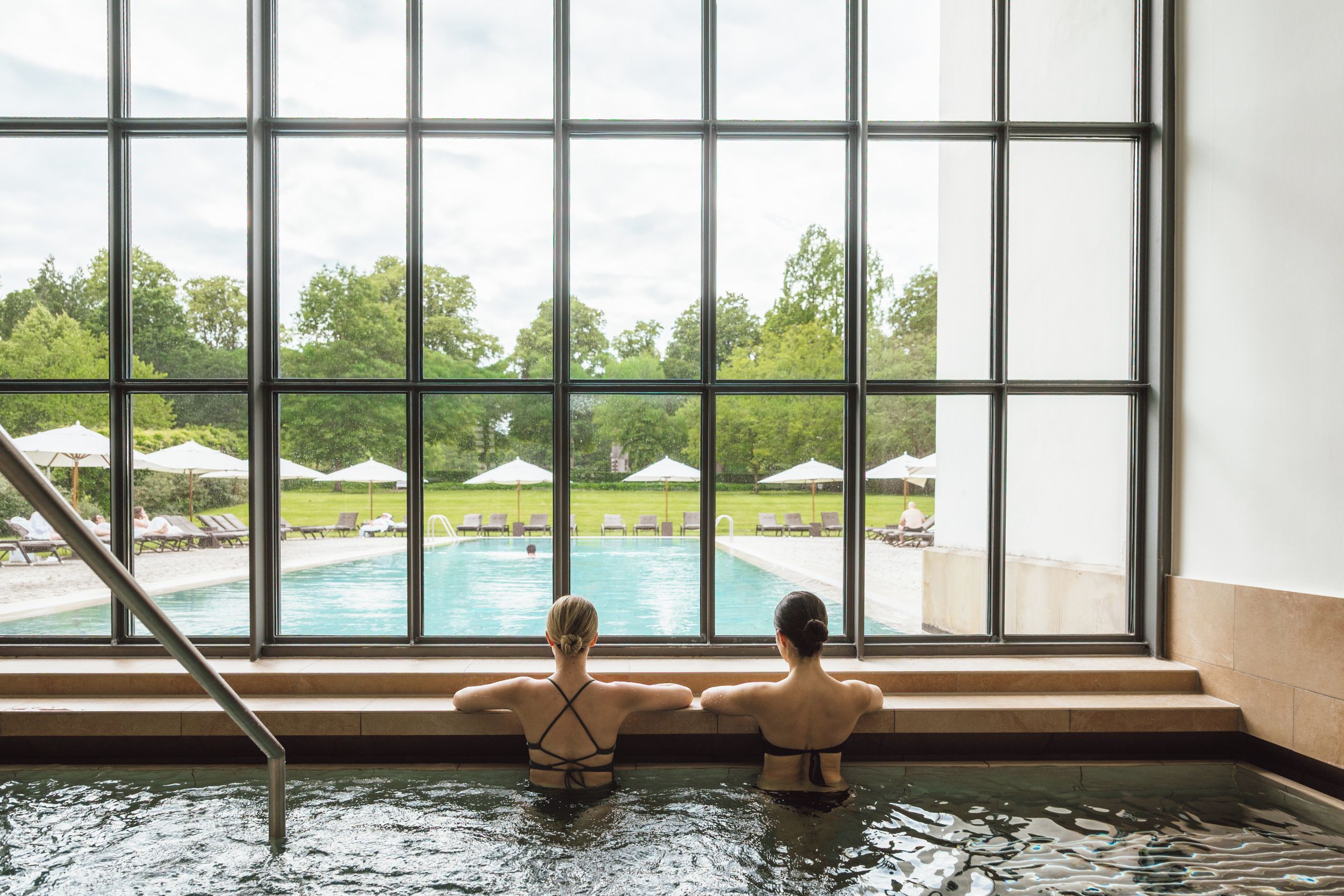 Two people in a swimming pool with a view of a poolside area and umbrellas.